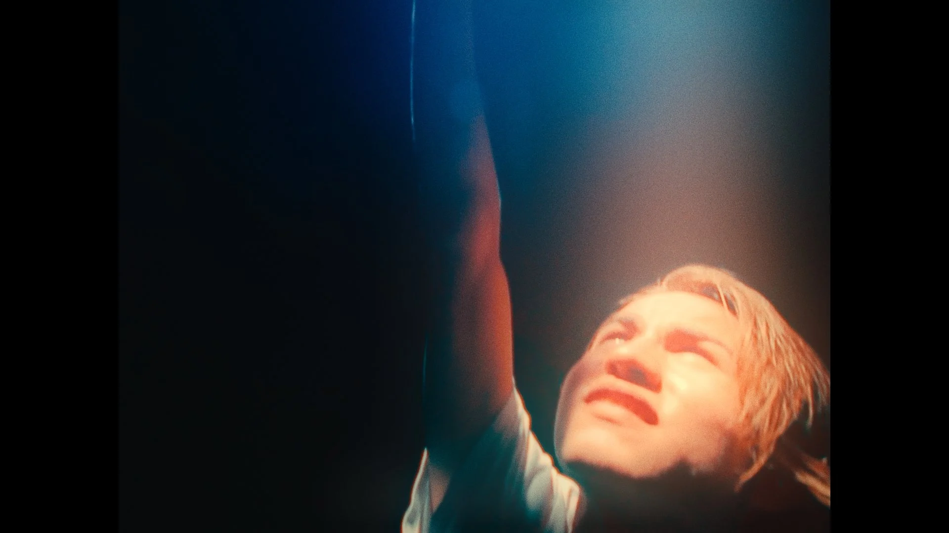 A young boy with blonde hair grimacing and closing his eyes, reaching upwards with his left hand, in a dimly lit setting with a spotlight shining down on him.