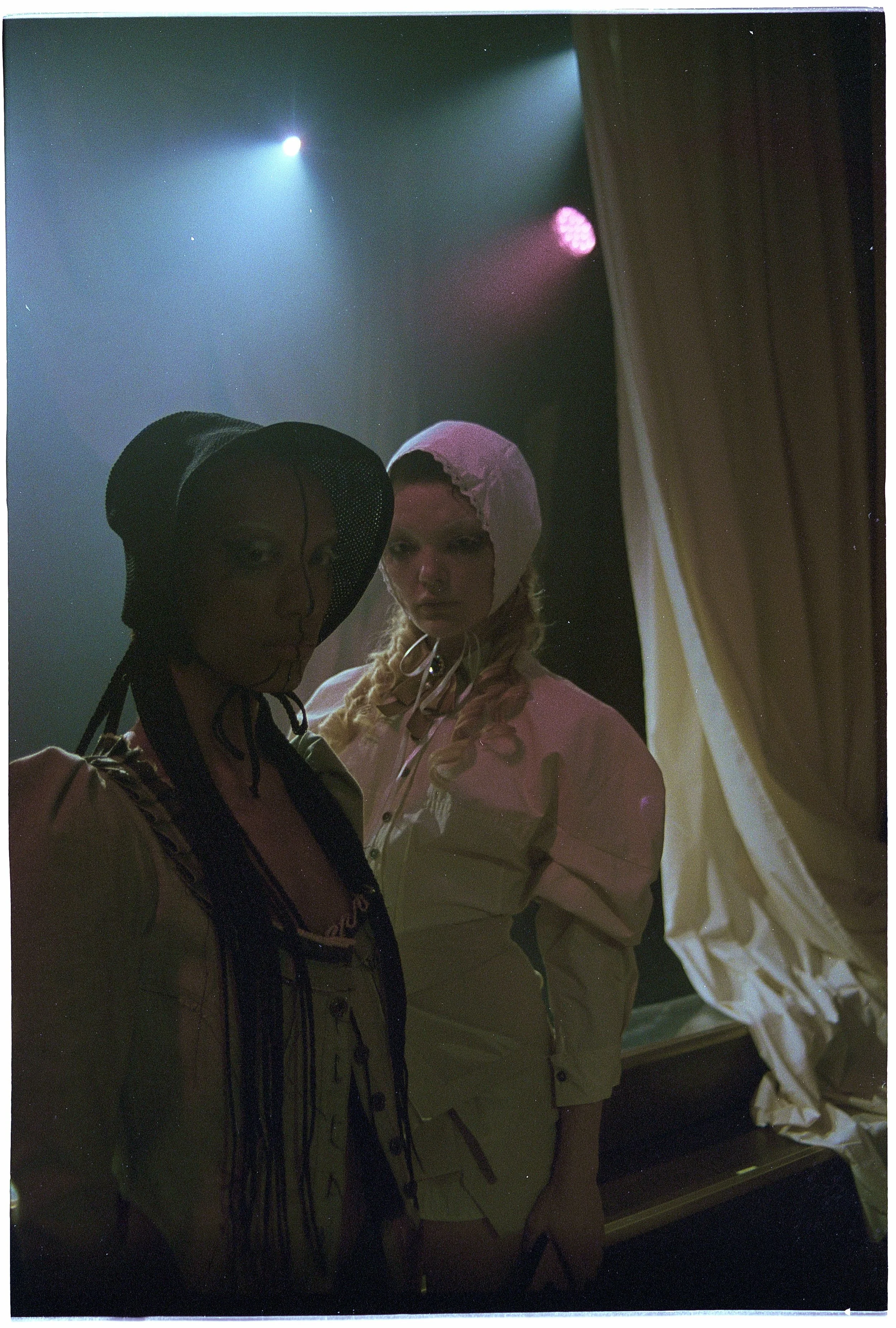 Two women in vintage clothing and bonnets stand behind a stage curtain, with colorful stage lights overhead.