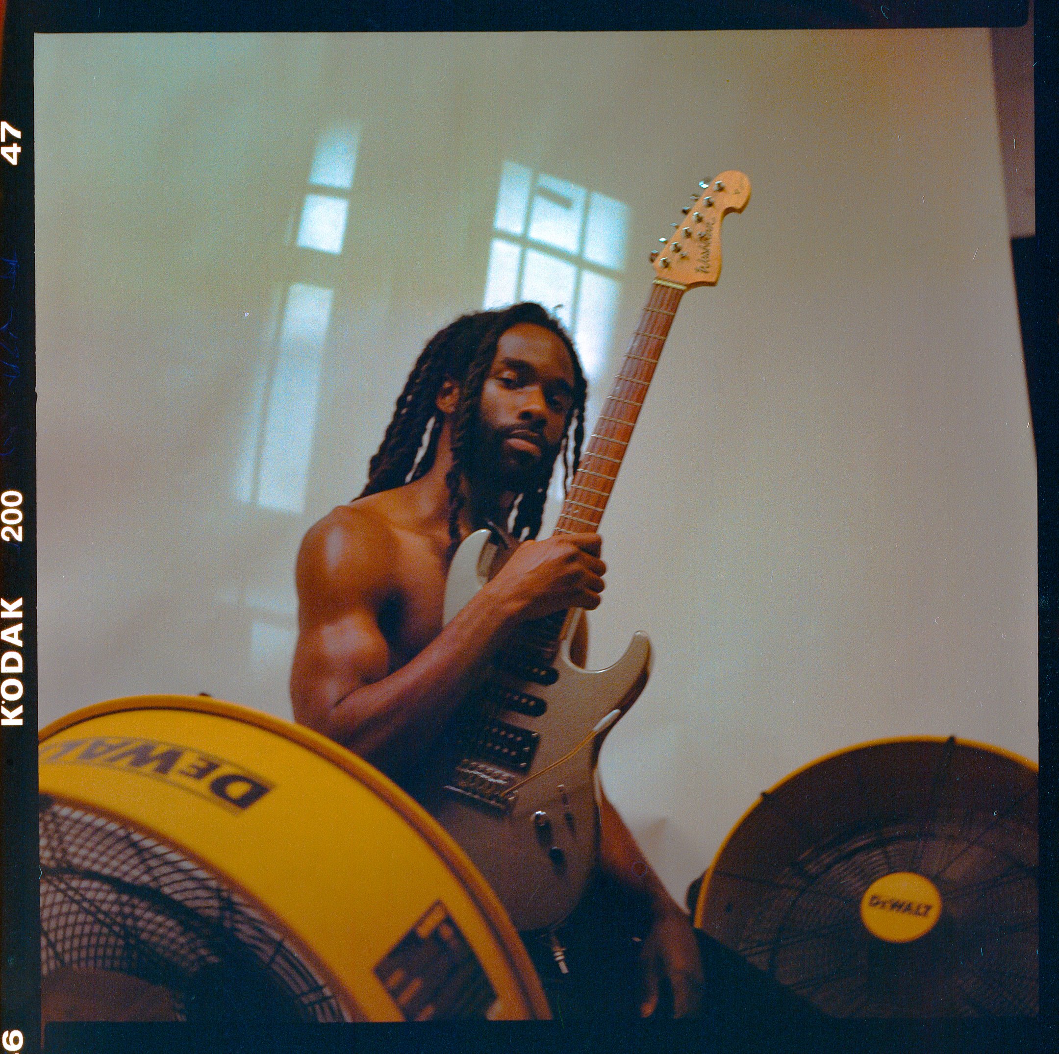 A shirtless man with dreadlocks playing an electric guitar, with two yellow fans on either side. The background is a light-colored wall with a window reflection.