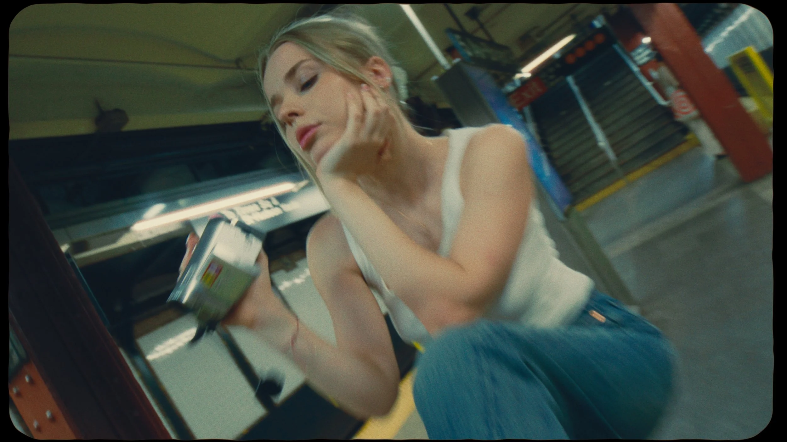 A young woman with blonde hair is sitting on the floor in a store aisle, looking at a smartphone she is holding in her right hand. She is wearing a white sleeveless top and blue jeans, with her left hand resting on her face, and appears to be deep in