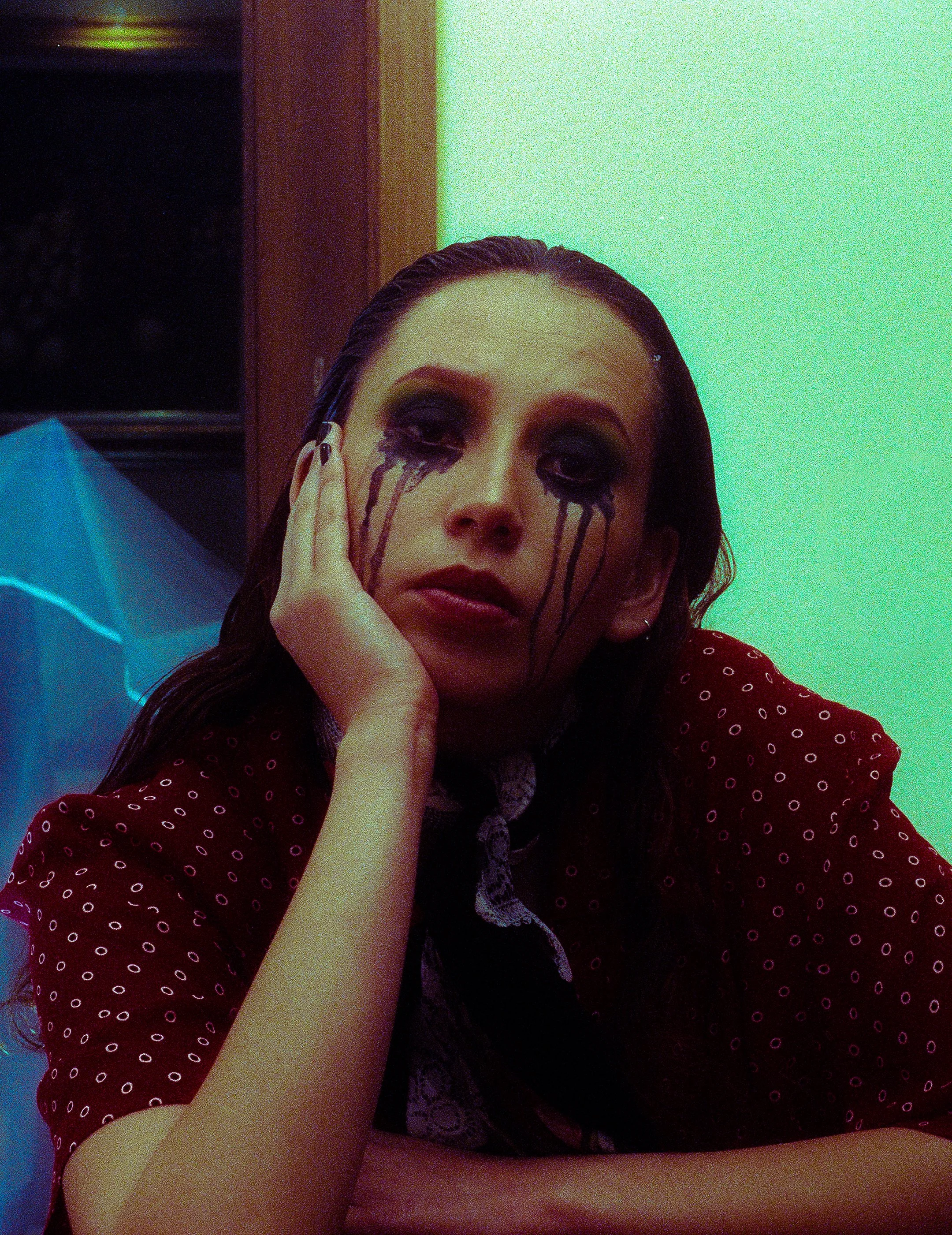 A woman with dark hair and makeup, wearing a red polka dot shirt, with black tears painted around her eyes, resting her face on her hand, sitting in a room with colorful lighting.