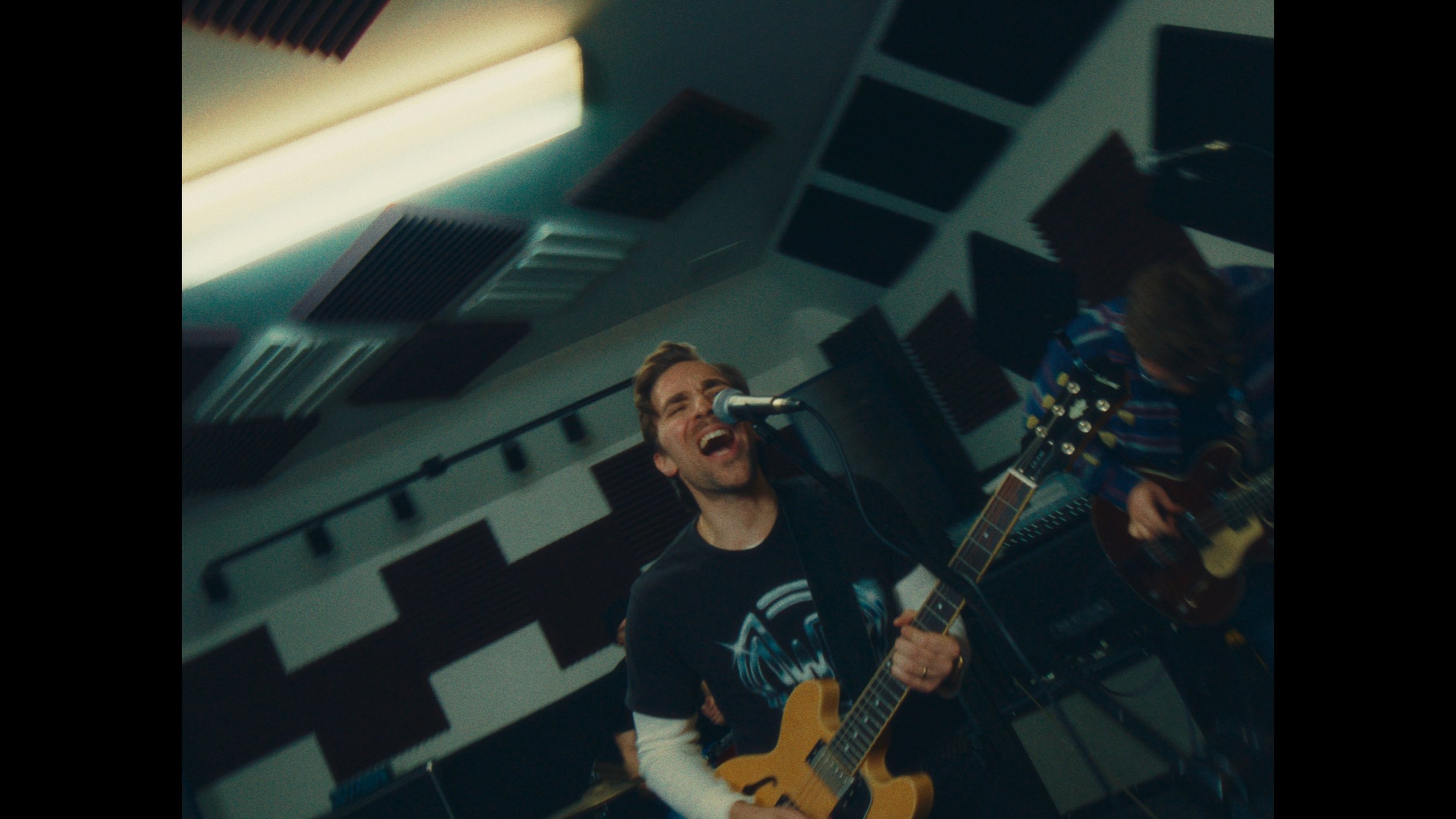 A man singing into a microphone while playing an electric guitar in a music studio, with another person playing an acoustic guitar in the background.