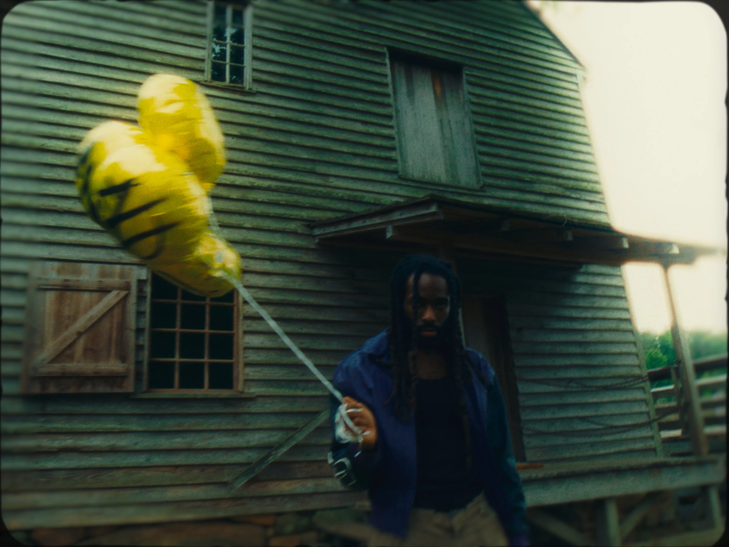 A person holding a yellow smiley face balloon in front of a wooden house with closed shutters.