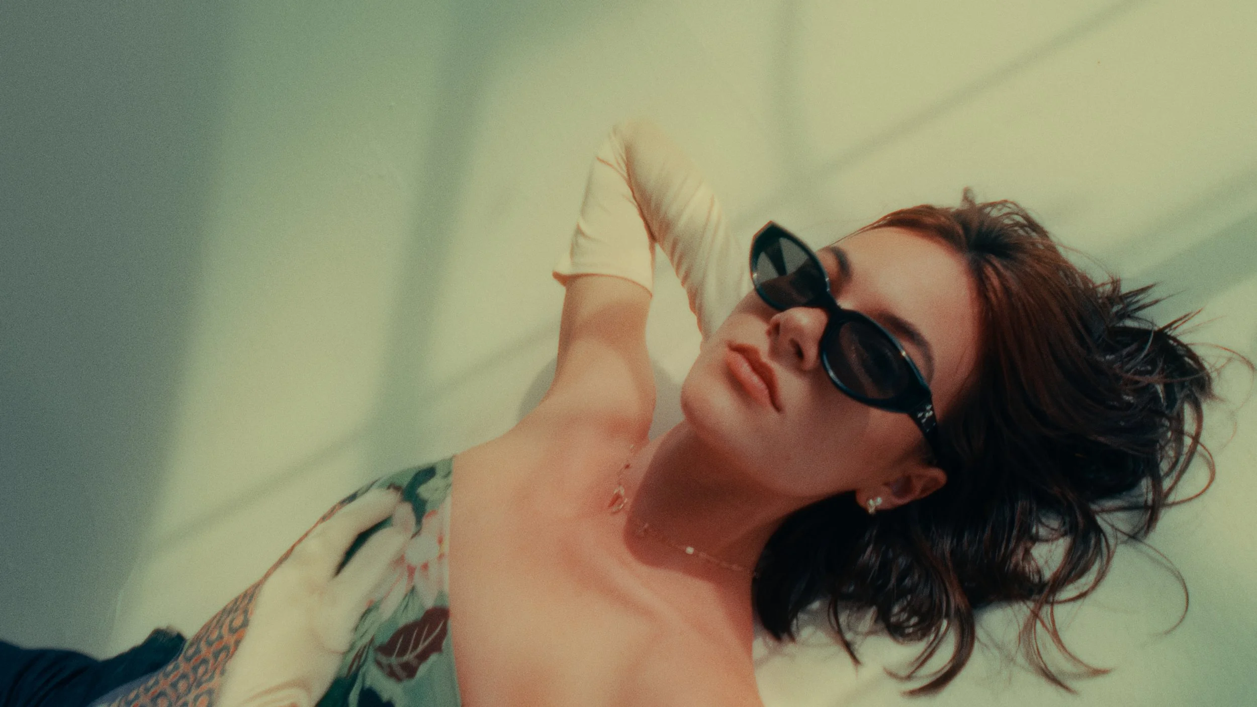 A woman with curly brown hair and black sunglasses lying on her side, wearing a patterned strapless top and a white glove on one arm.