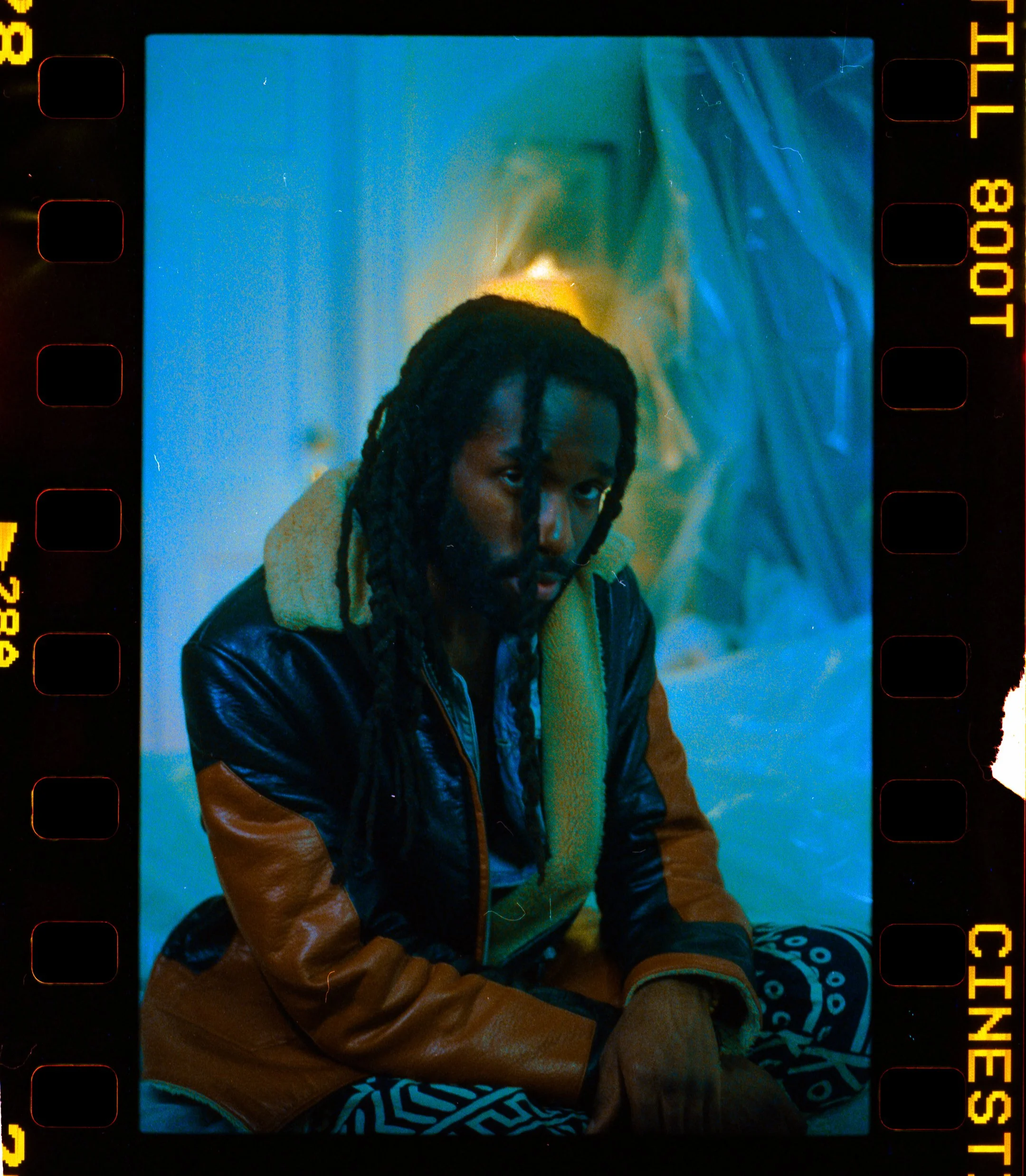 A man with dreadlocks and a beard sitting indoors, wearing a leather jacket with a shearling collar, patterned pants, and gloves, with a serious expression, illuminated by blue and warm lighting.