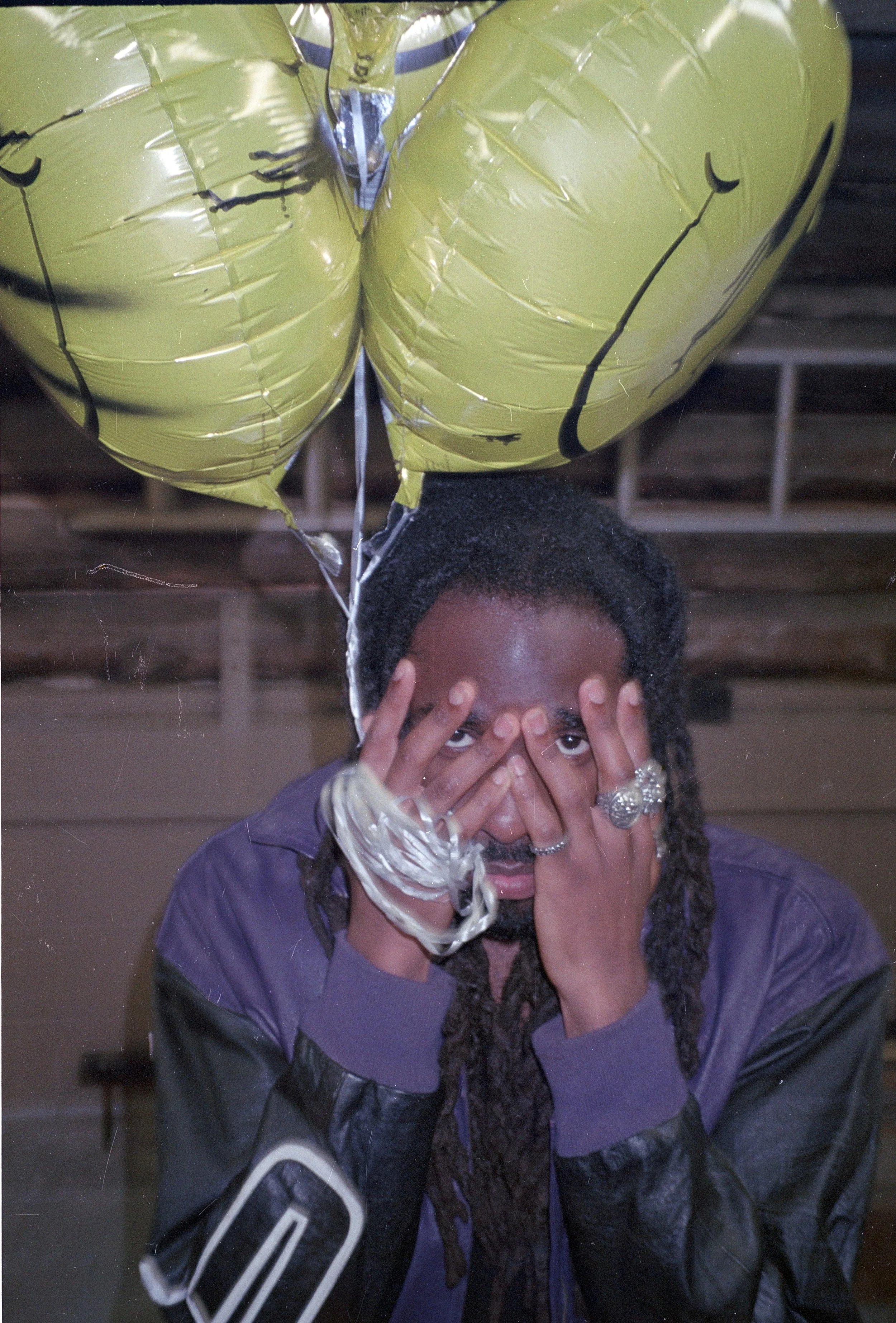 A person with dreadlocks covering their face with both hands, wearing multiple rings and rings on their fingers. They are holding two large yellow balloons with cartoonish faces, and the background is a wooden ceiling inside a building.