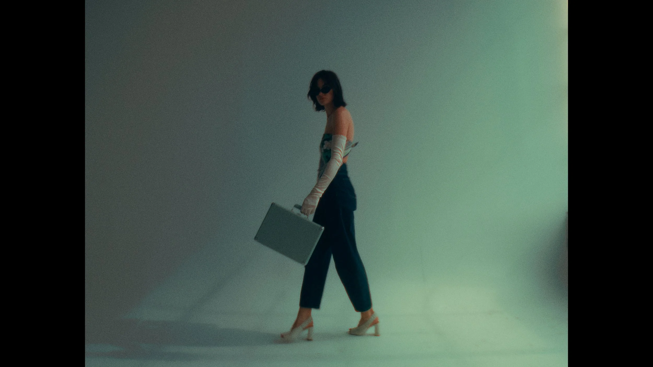 A woman with dark hair, wearing sunglasses, a strapless top, long gloves, high heels, and dark pants, is holding a briefcase while walking in a plain, gradient background.