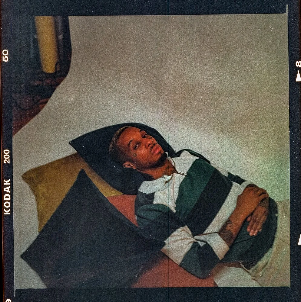 A young man with tattoos, lying on a bed with pillows, looking at the camera. The wall behind him is plain and light-colored.