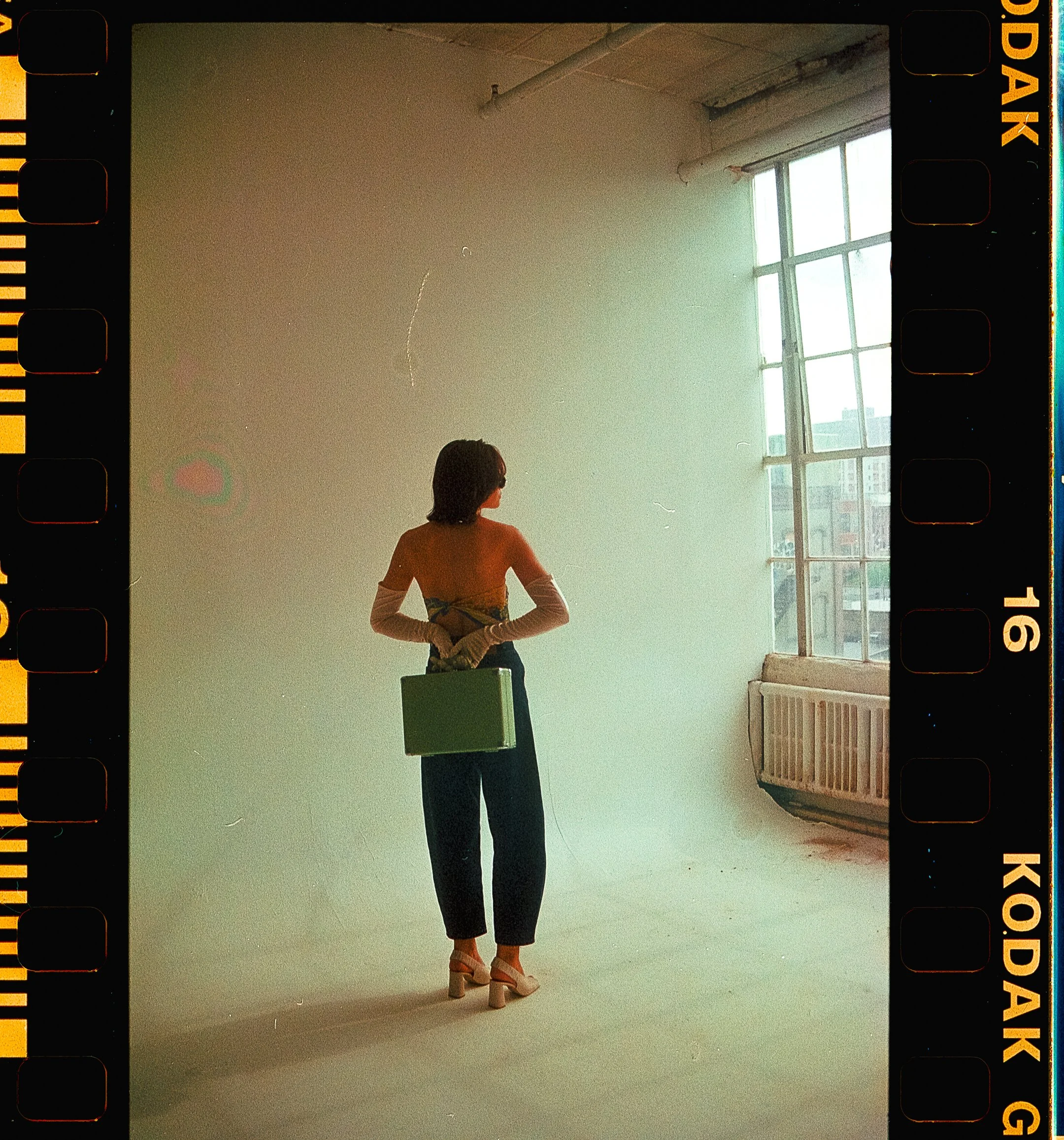 A woman with shoulder-length dark hair standing in front of a plain white wall, holding a green briefcase in her right hand, wearing high-heeled shoes, dark pants, and an off-shoulder top. She is looking out the large window on her right, which lets in natural light and shows a cityscape outside.