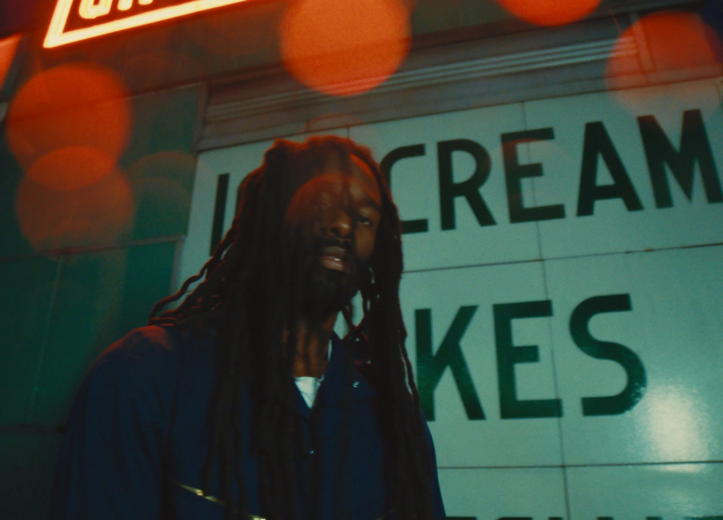 A man with long dreadlocks standing in front of a sign that says "Ice Cream" and "Bakes," with orange and red bokeh lights in the background.