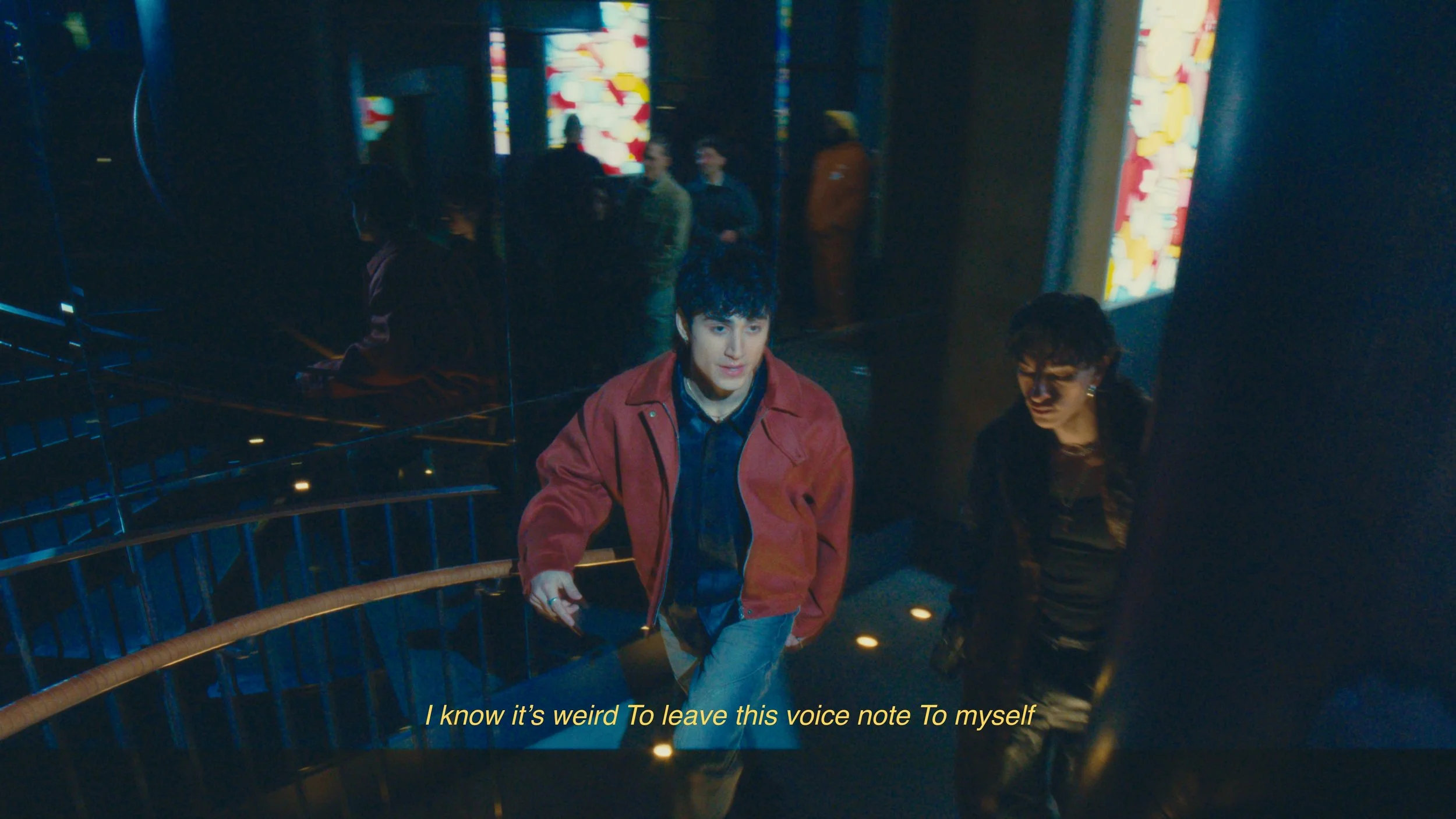 Two young people, one wearing a red jacket and the other in dark clothing, walking up a staircase inside a modern building with colorful abstract artwork on the walls. The scene has dim lighting and a reflective wall.