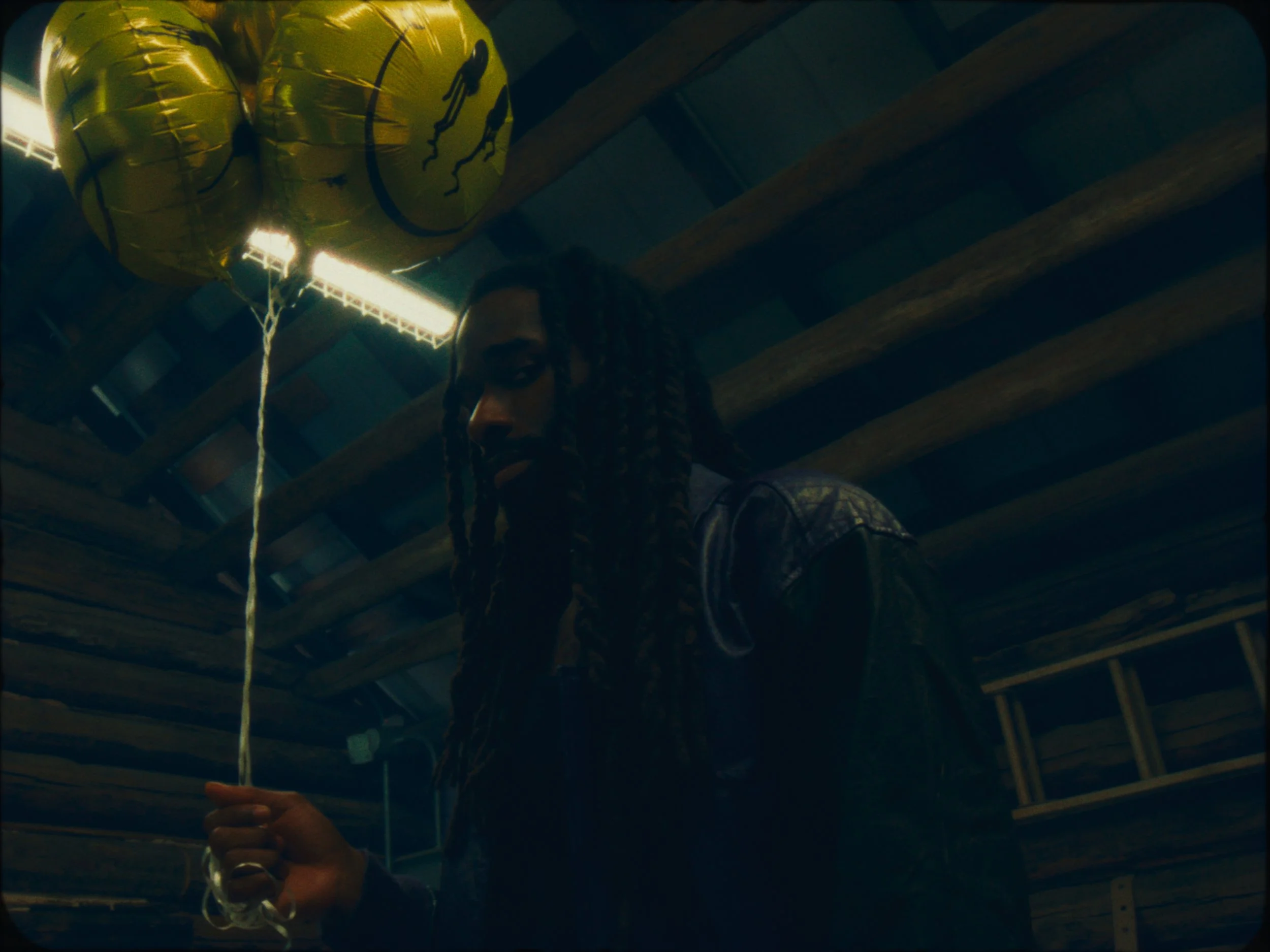 A person with long braided hair holding a gold balloon with a warning symbol in a dimly lit, rustic setting.