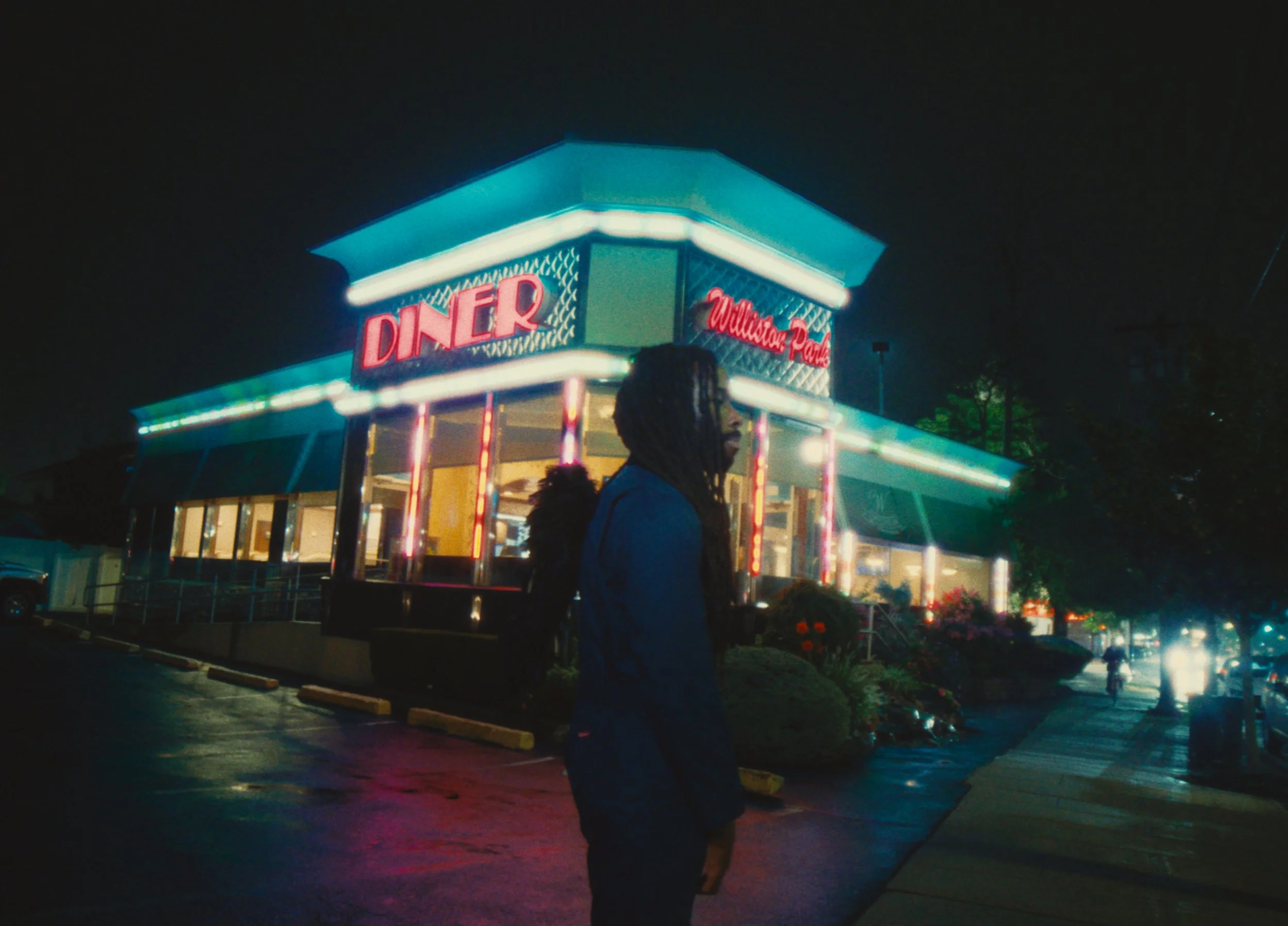 Nighttime scene outside a retro diner with neon lights, a woman with dreadlocks in the foreground, and cars on the street.