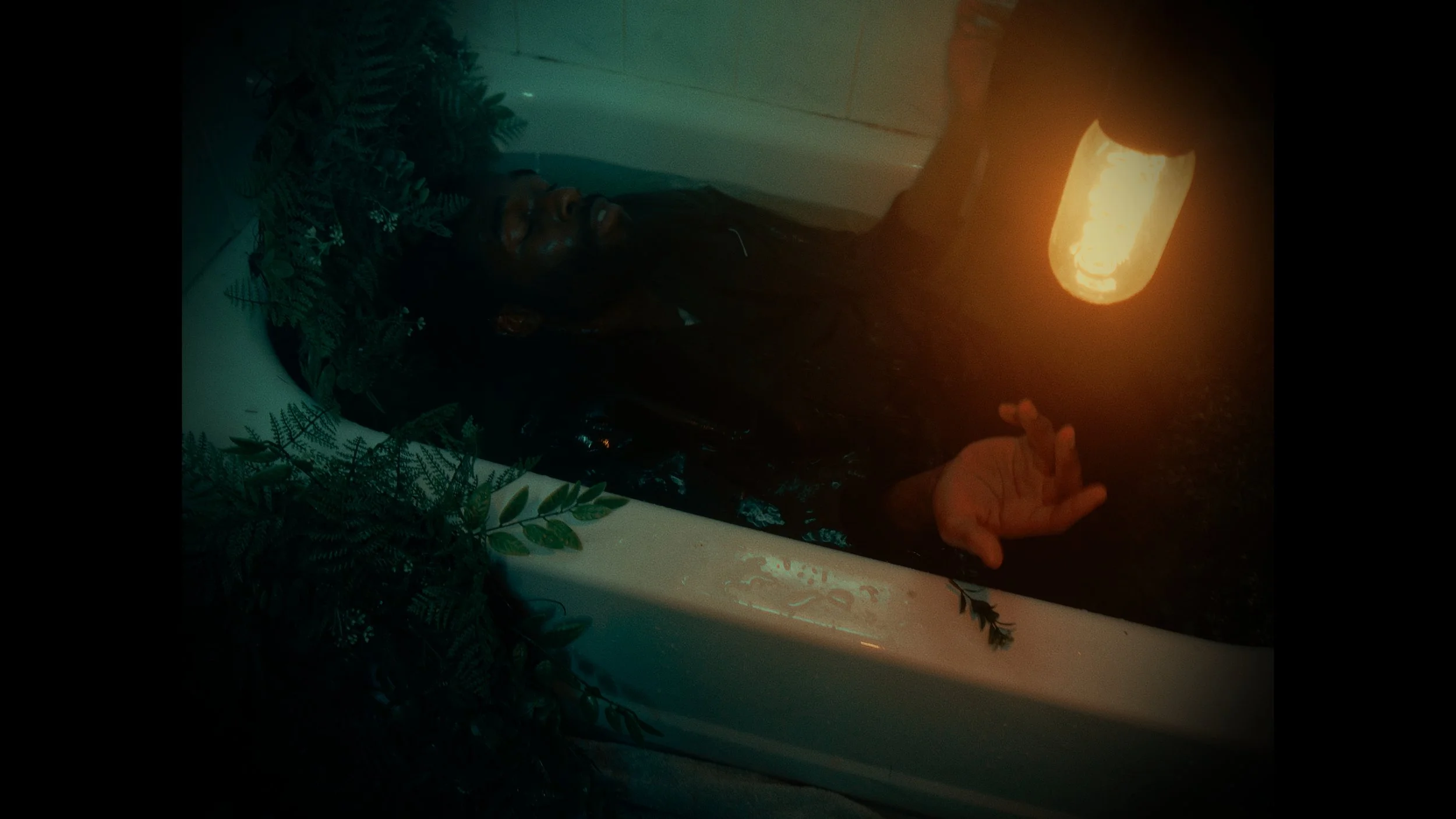 A person with long dark hair lying in a bathtub filled with water, holding a lantern that illuminates their face and hand, with greenery around the bathtub.