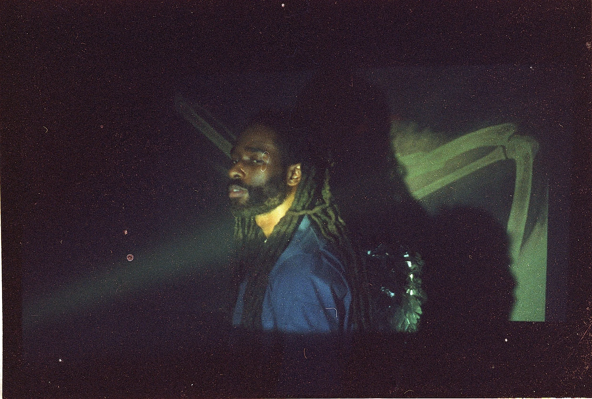 A man with dreadlocks, glasses, and a beard sitting inside a car at night with a dark background.