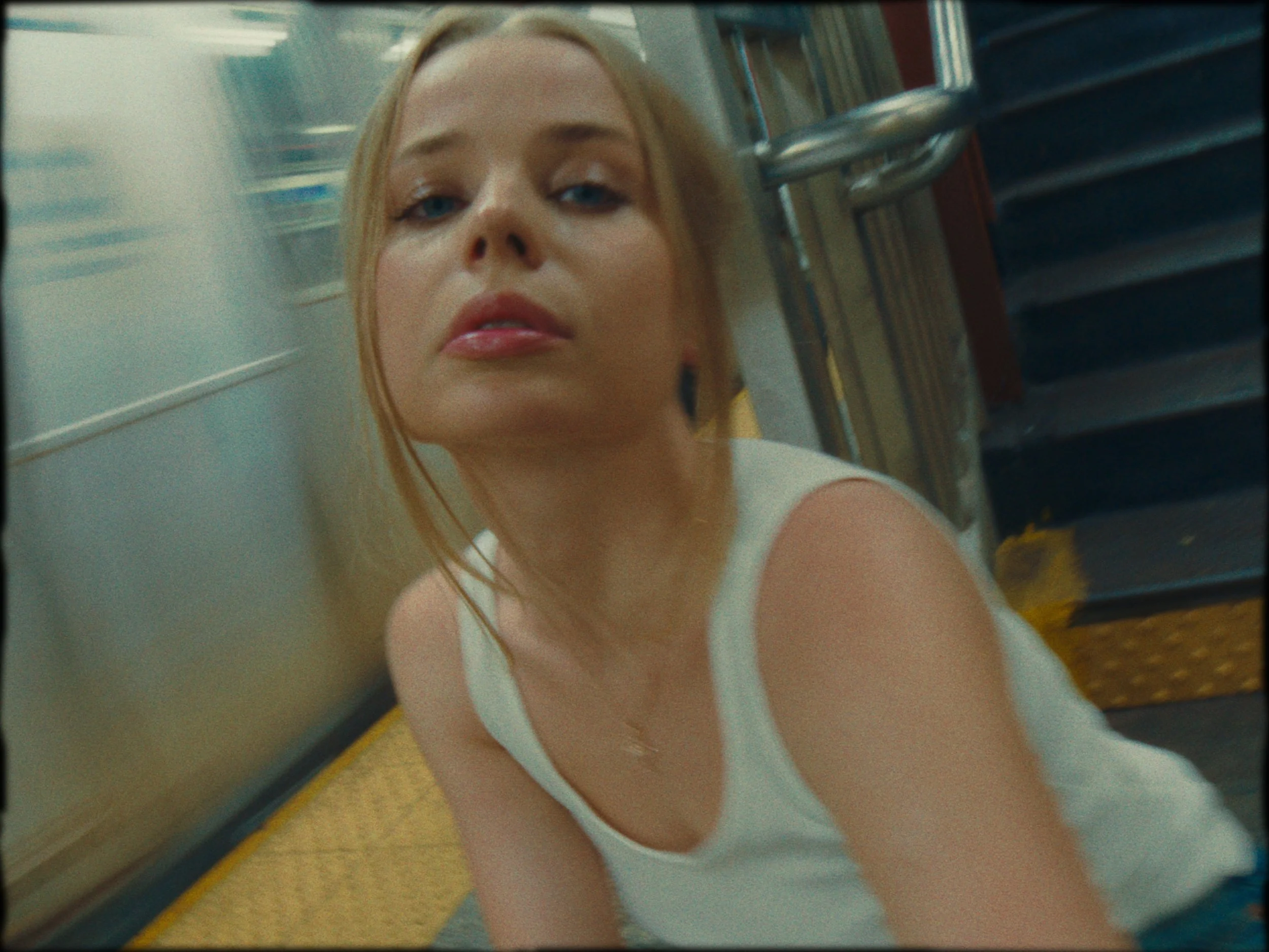 A woman with light skin and blonde hair looking up while leaning on the floor of a subway or train, with stairs and a metallic handrail visible in the background.