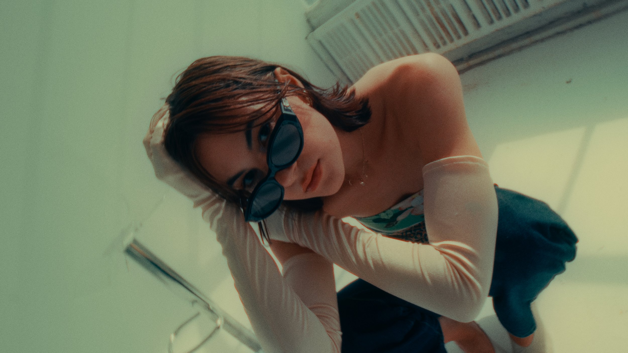 A young woman with short brown hair, wearing sunglasses and a patterned sleeveless top, is sitting on the floor and looking up at the camera, with a hand behind her head.