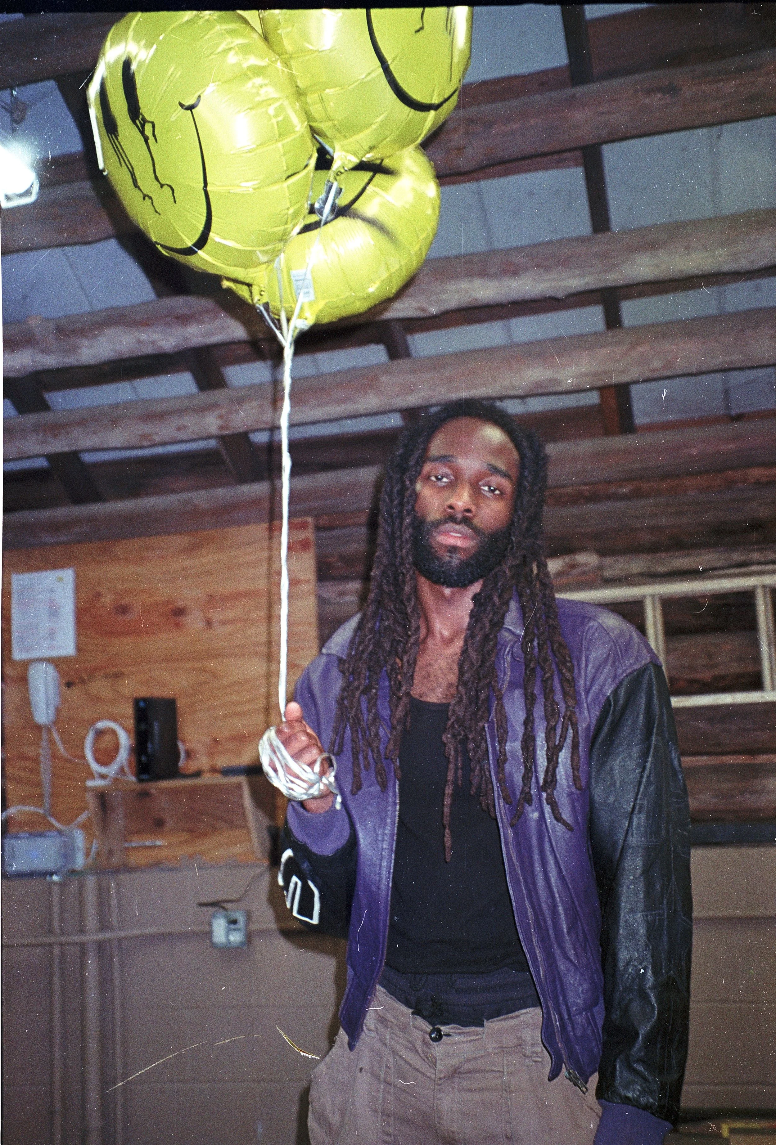 A man with dreadlocks and a beard holding a yellow smiley face helium balloon, standing in a wooden interior space with exposed beams.