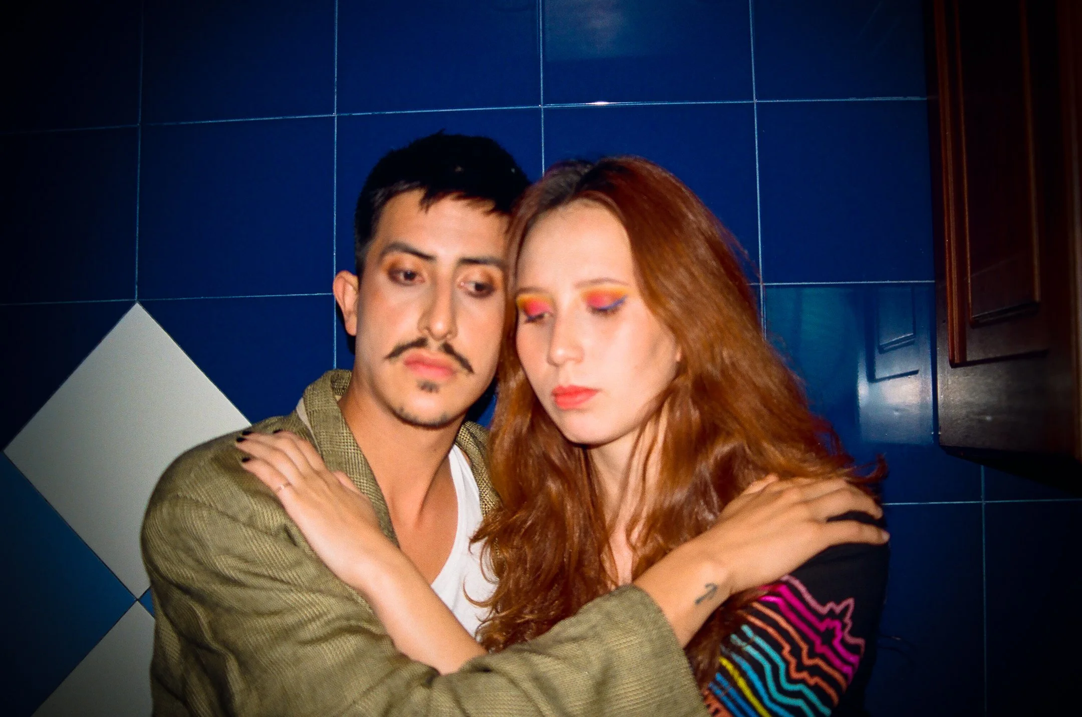 A man and a woman embrace each other, with both having their arms around each other's shoulders. The man has dark hair, a mustache, and a beard, and is wearing a beige jacket over a white shirt. The woman has long reddish hair with colorful eye makeup, and is wearing a black top with colorful patterns on the sleeves. They are standing against a dark blue tiled wall with some white tiles and a wooden cabinet on the right.