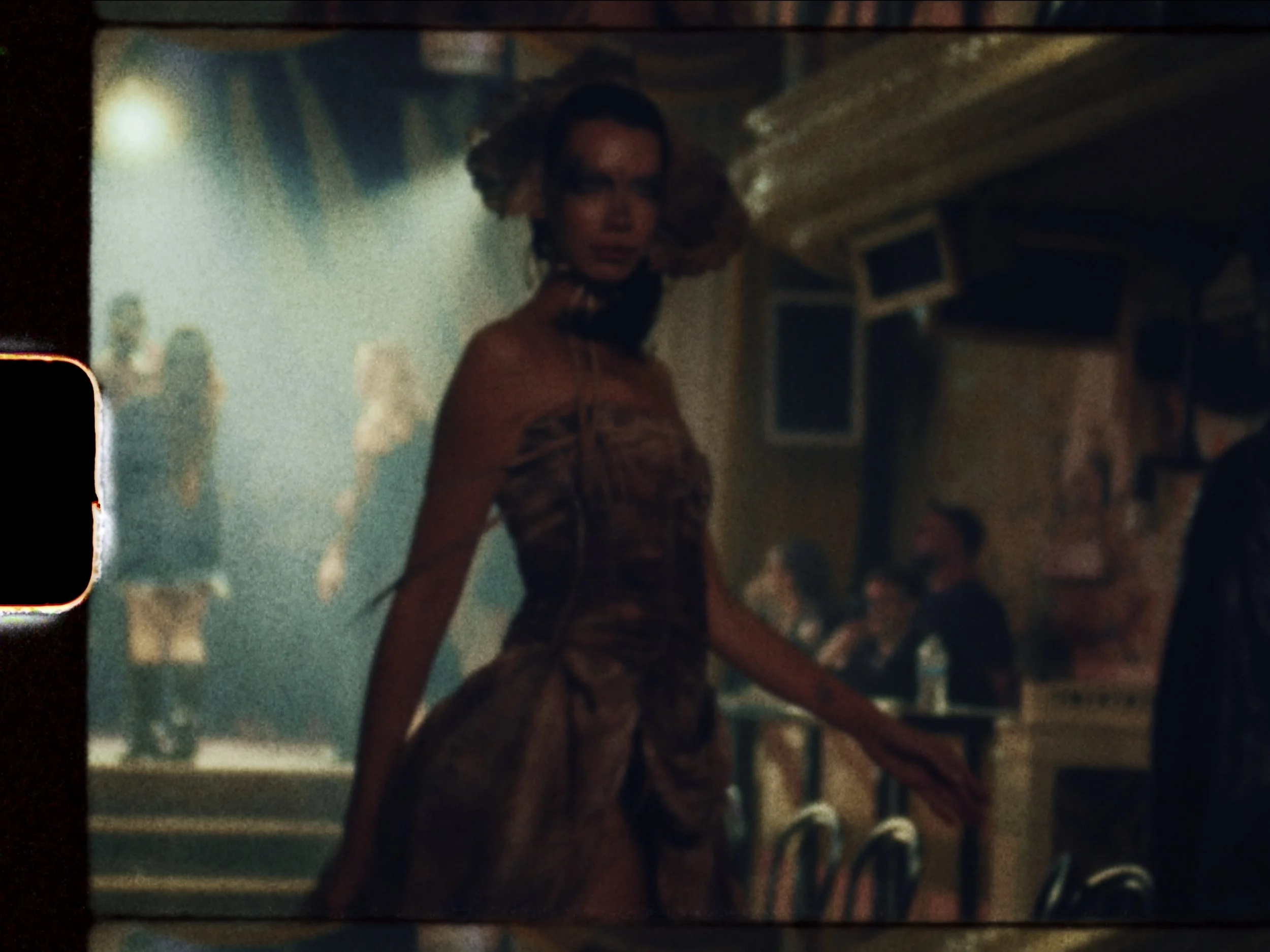 A woman with curly blonde hair and glasses in a vintage dress is in a dimly lit social setting, possibly a bar or club, with other people in the background.