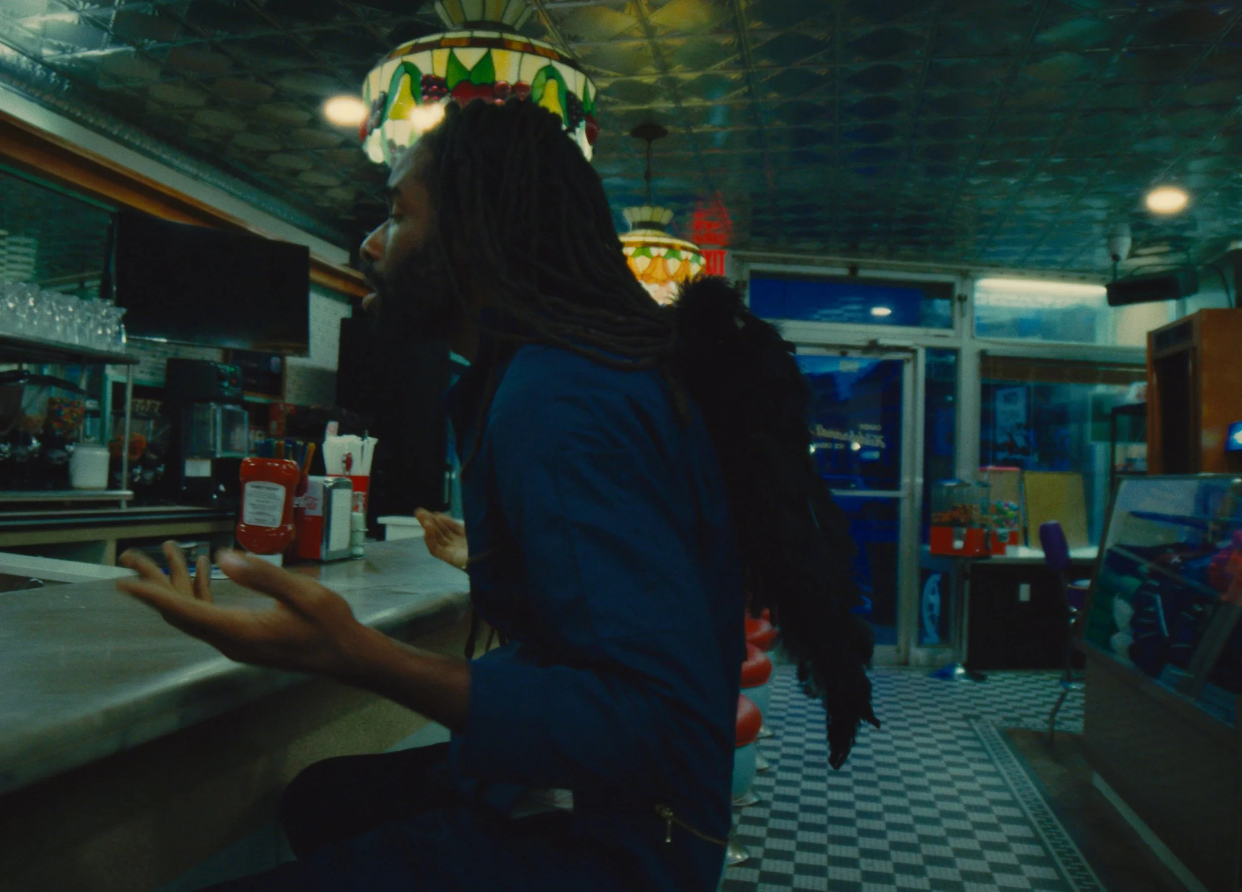 A man with long dreadlocks and a blue jacket sitting at a counter inside a diner or cafe, talking to someone off-camera. The background includes a colorful stained glass lamp, red ketchup bottles, and a checkered black and white floor.