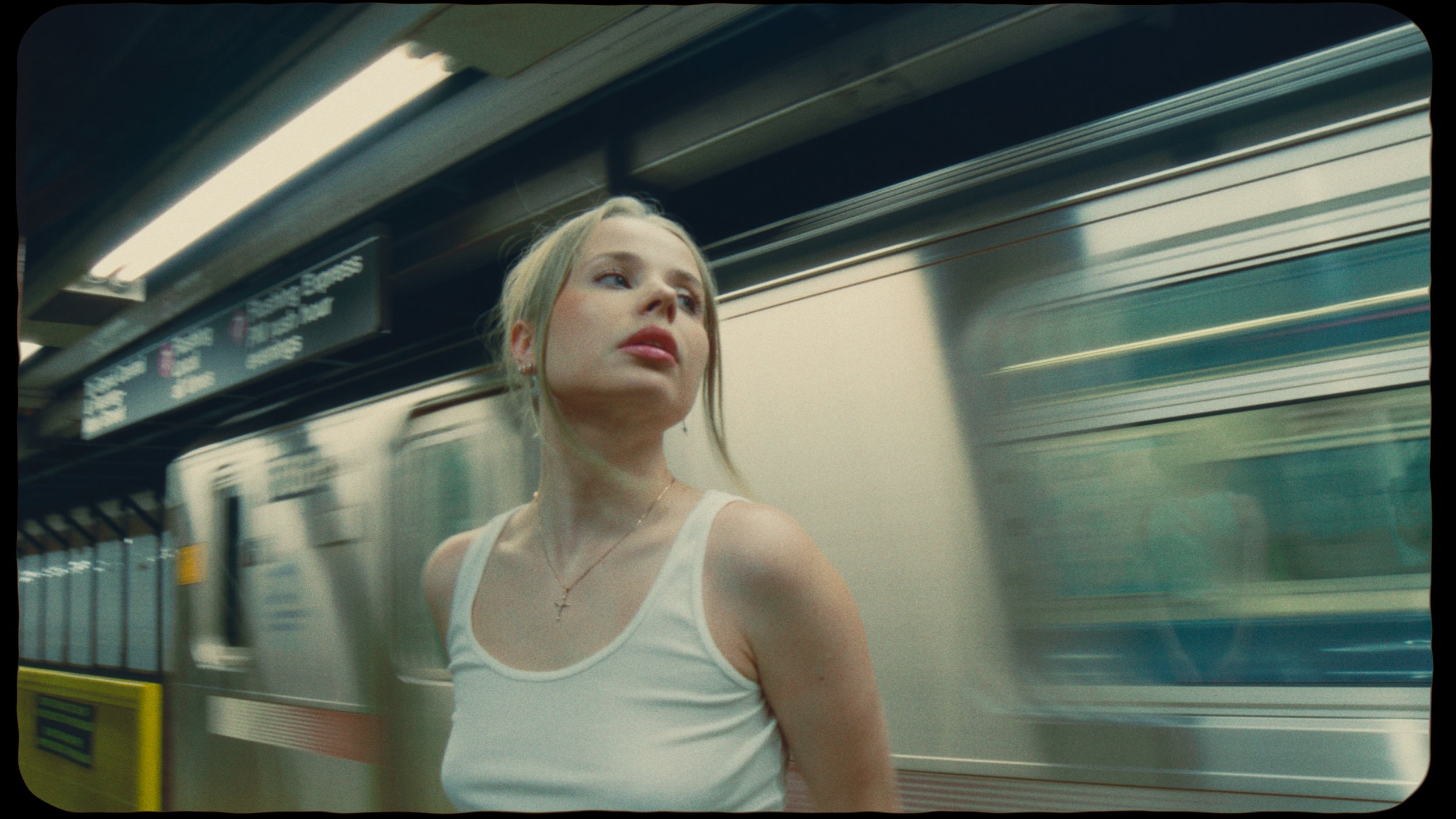 A young woman with blonde hair, wearing a white tank top and a cross necklace, stands on a subway platform with a moving train in the background.