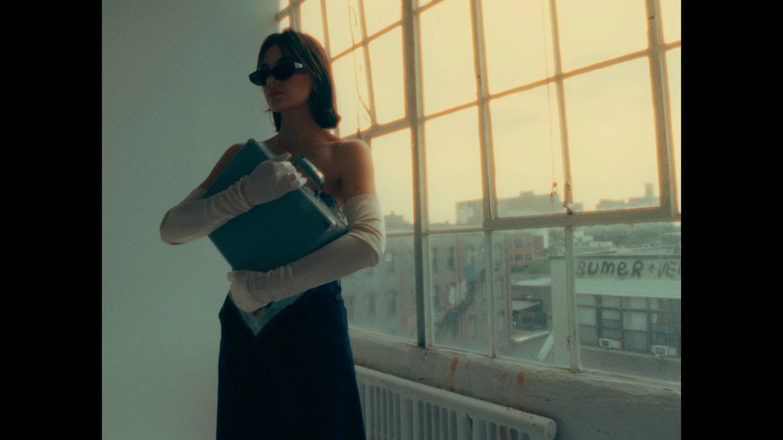 A woman with shoulder-length brown hair wearing sunglasses, white gloves, a strapless top, and dark pants, holding a blue binder and a book, standing in front of a large window with a cityscape view at sunset.