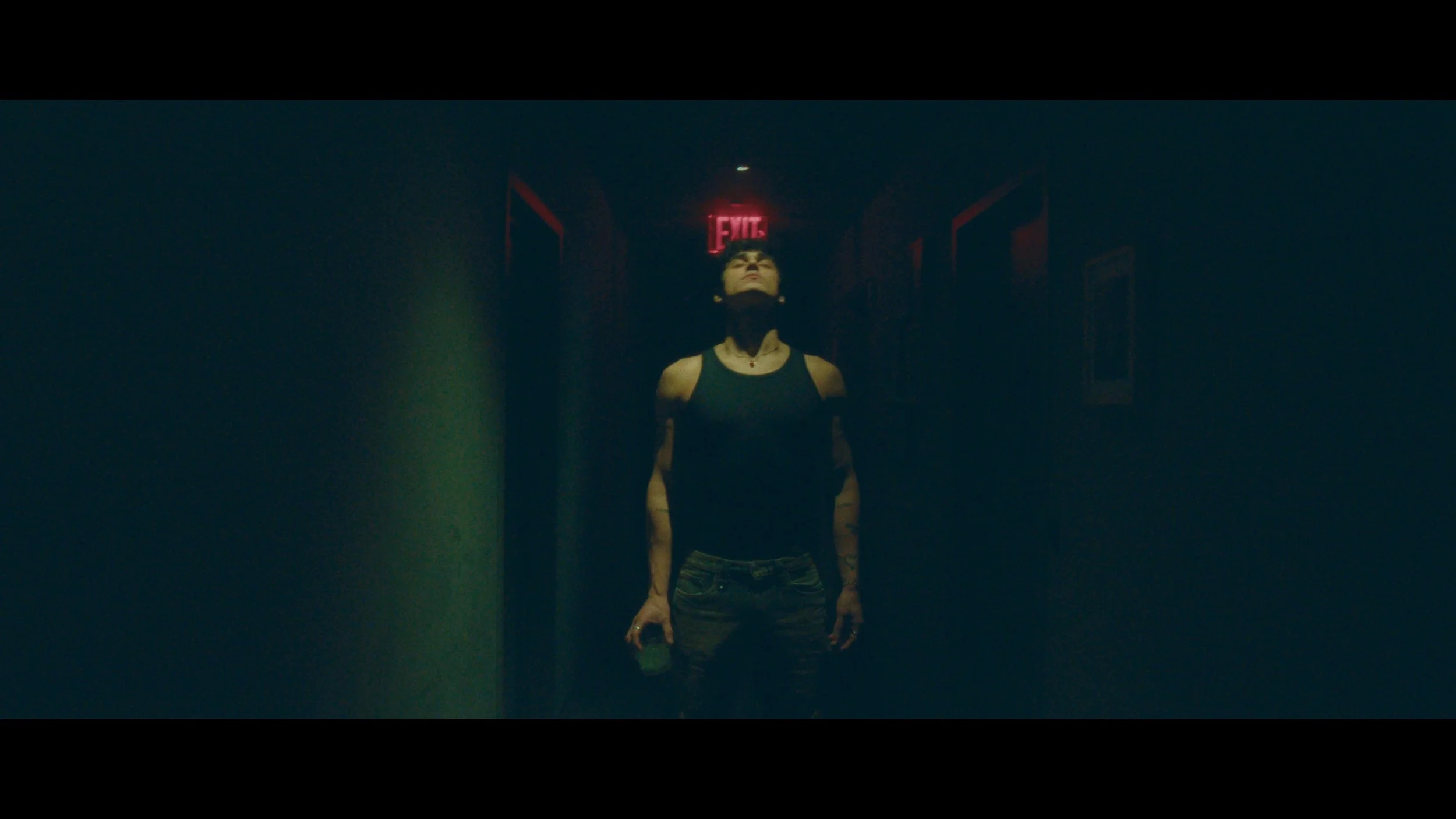 A woman standing in a dimly lit corridor with a red exit sign above her head, looking upward, holding a small object in her right hand.