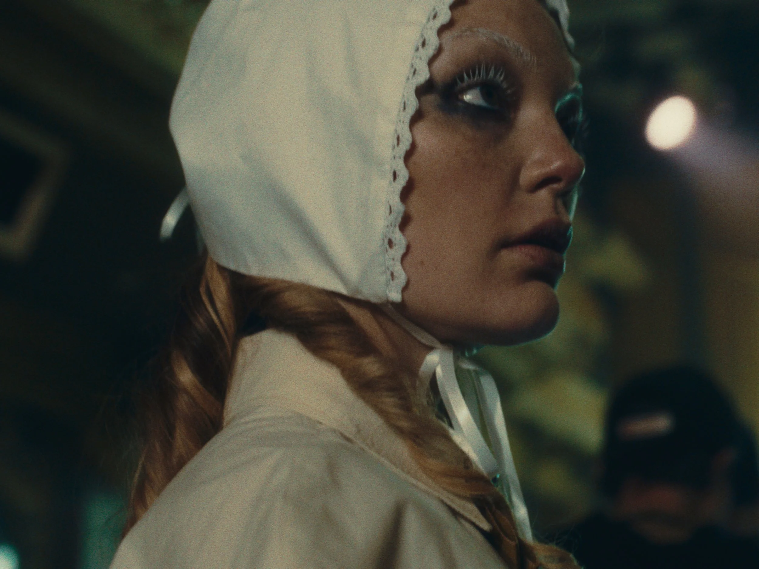 A woman with braided hair and makeup wearing a white lace hooded top, looking to the side in an outdoor setting at night.