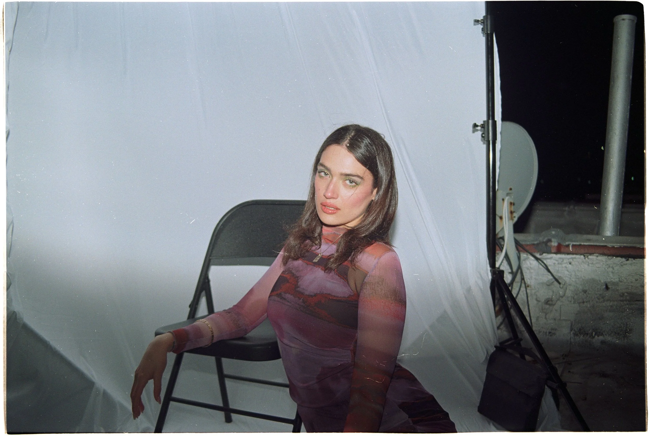 A woman with long dark hair, wearing a sheer, colorful, patterned top, sitting on a black chair in front of a white backdrop, possibly during a photoshoot at night.