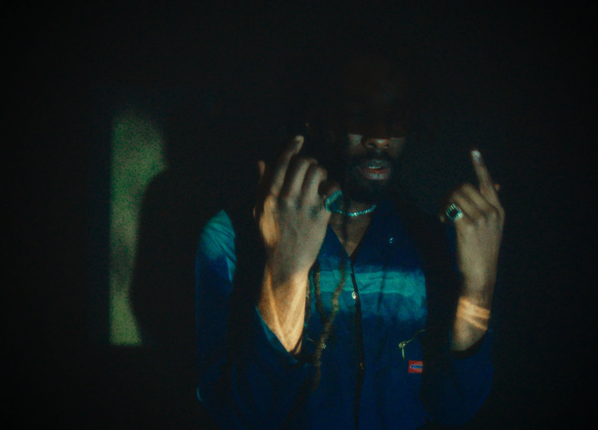 A person with dark hair and dark sunglasses adjusting their collar in a dimly lit environment.