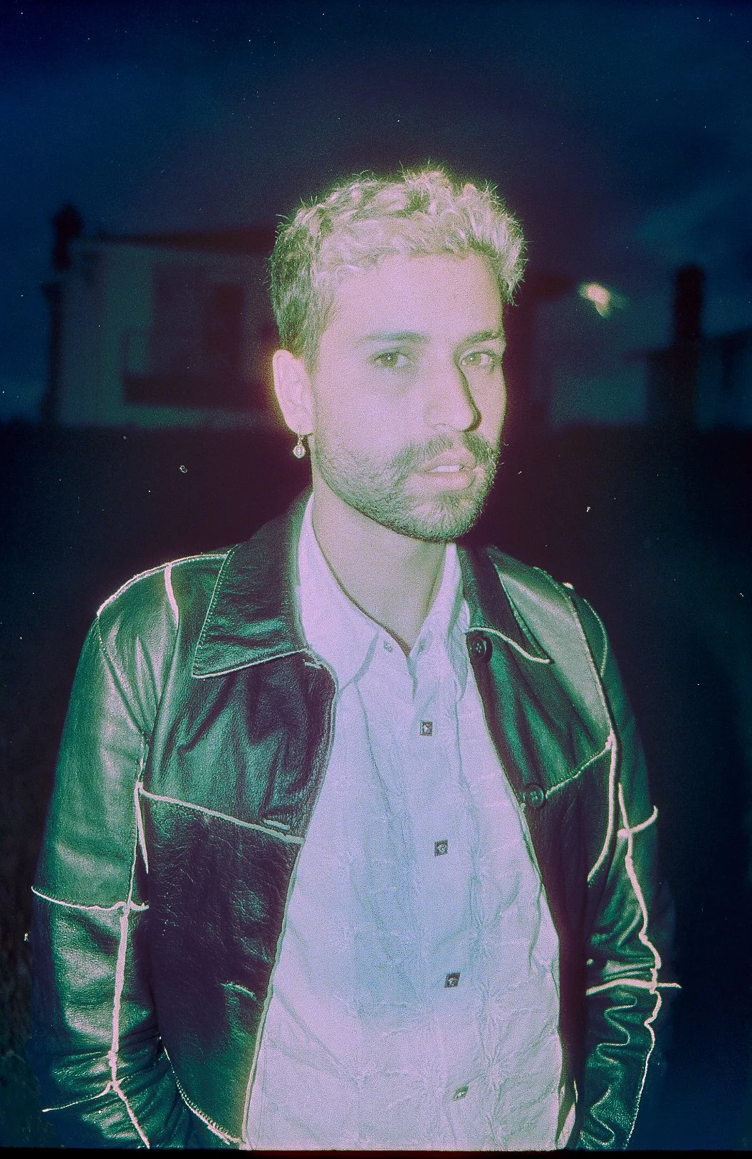 A man with a beard and short hair, wearing a leather jacket and white shirt, stands outdoors at night with a building and chimney in the background.