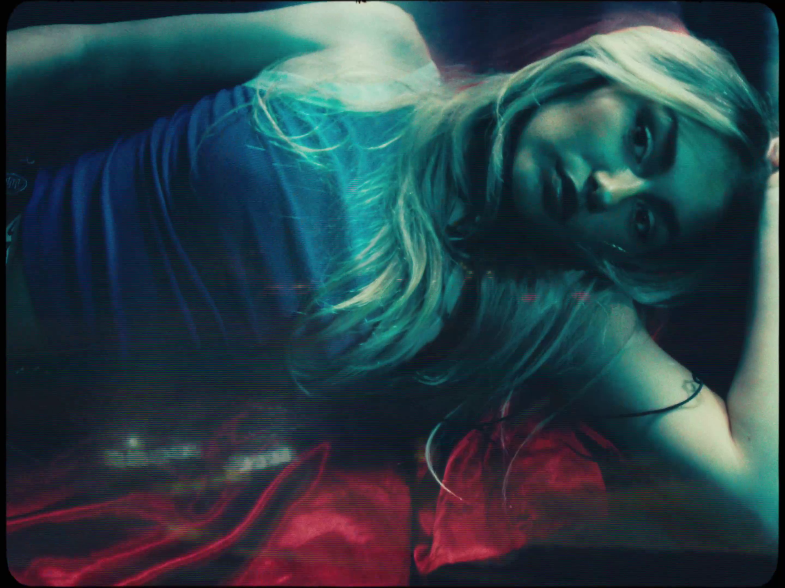 A woman with long blonde hair lying on her side on a bed with a red blanket, looking at the camera with a neutral expression.