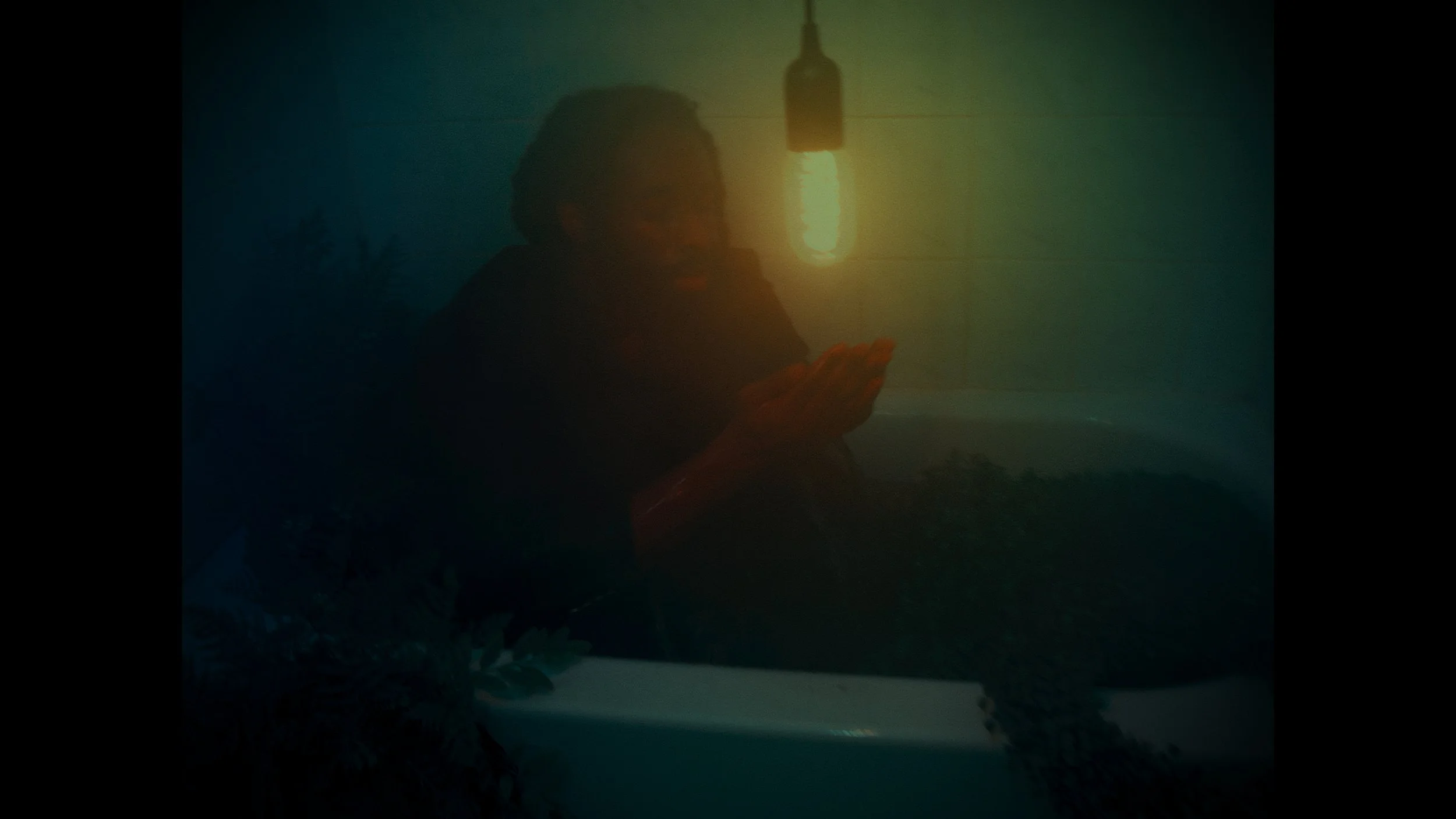 A person sitting in a dimly lit room with a single exposed lightbulb hanging from the ceiling, appearing to look at an object in their hands.