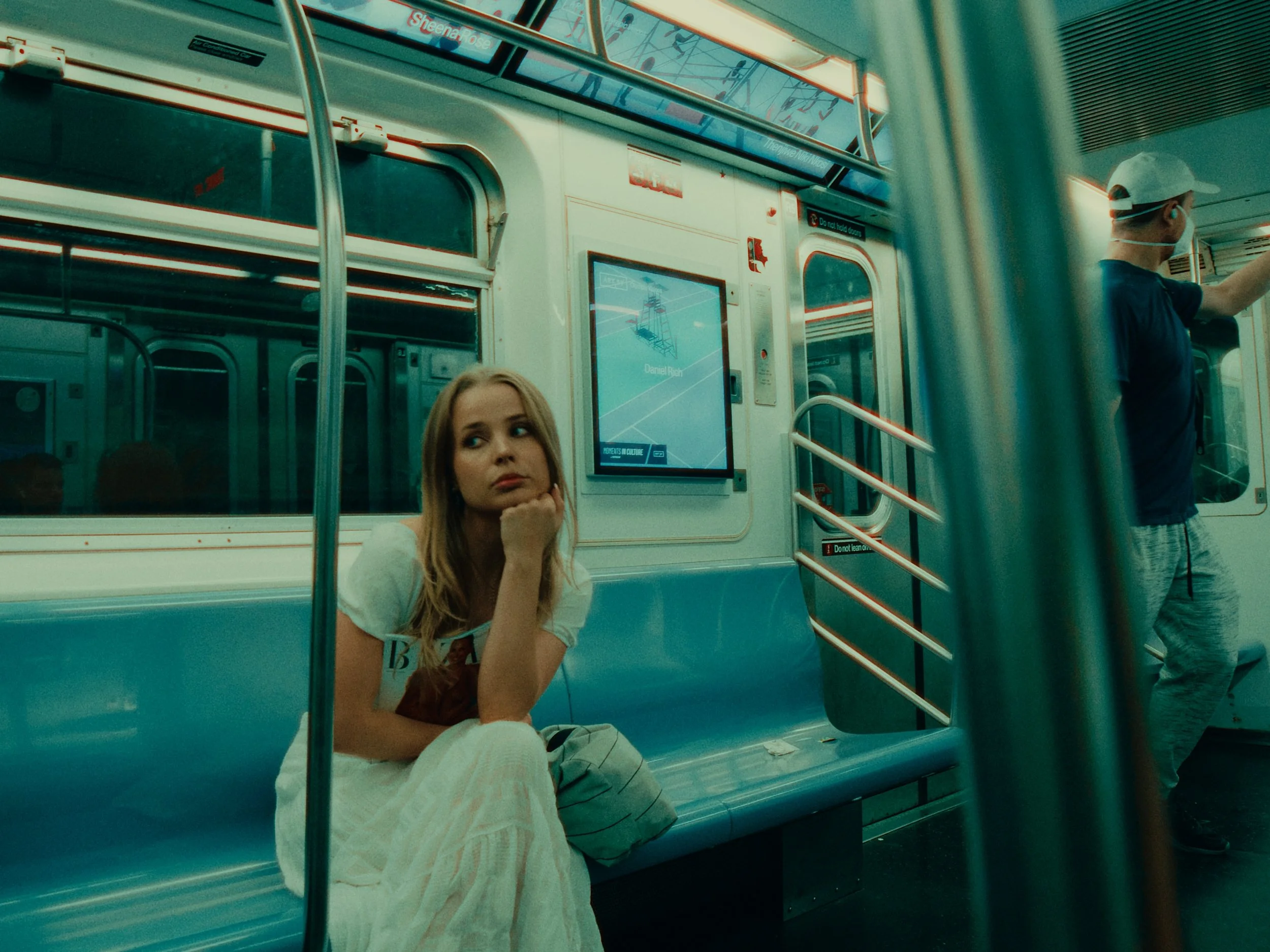 A woman sitting alone on a light blue subway train, looking thoughtfully towards the window, with a young man standing nearby wearing a face mask and cap, in an underground station.