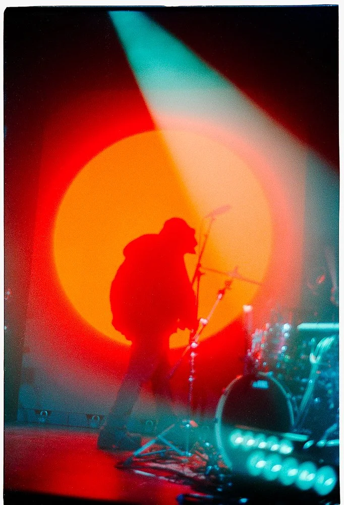 Musician performing on stage with silhouette backlit by large orange and red sun projection, surrounded by stage lights and equipment.