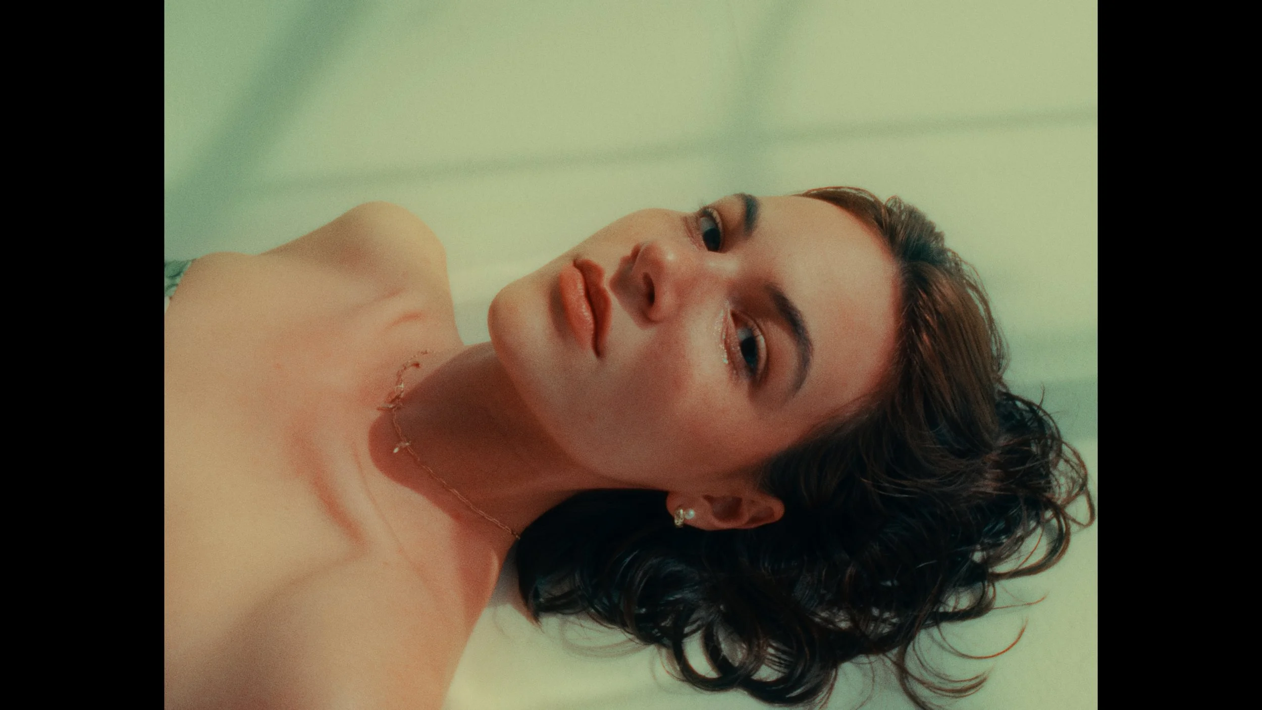 A woman with curly brown hair and earrings lies on her back on a light-colored surface, looking at the camera with a relaxed expression.