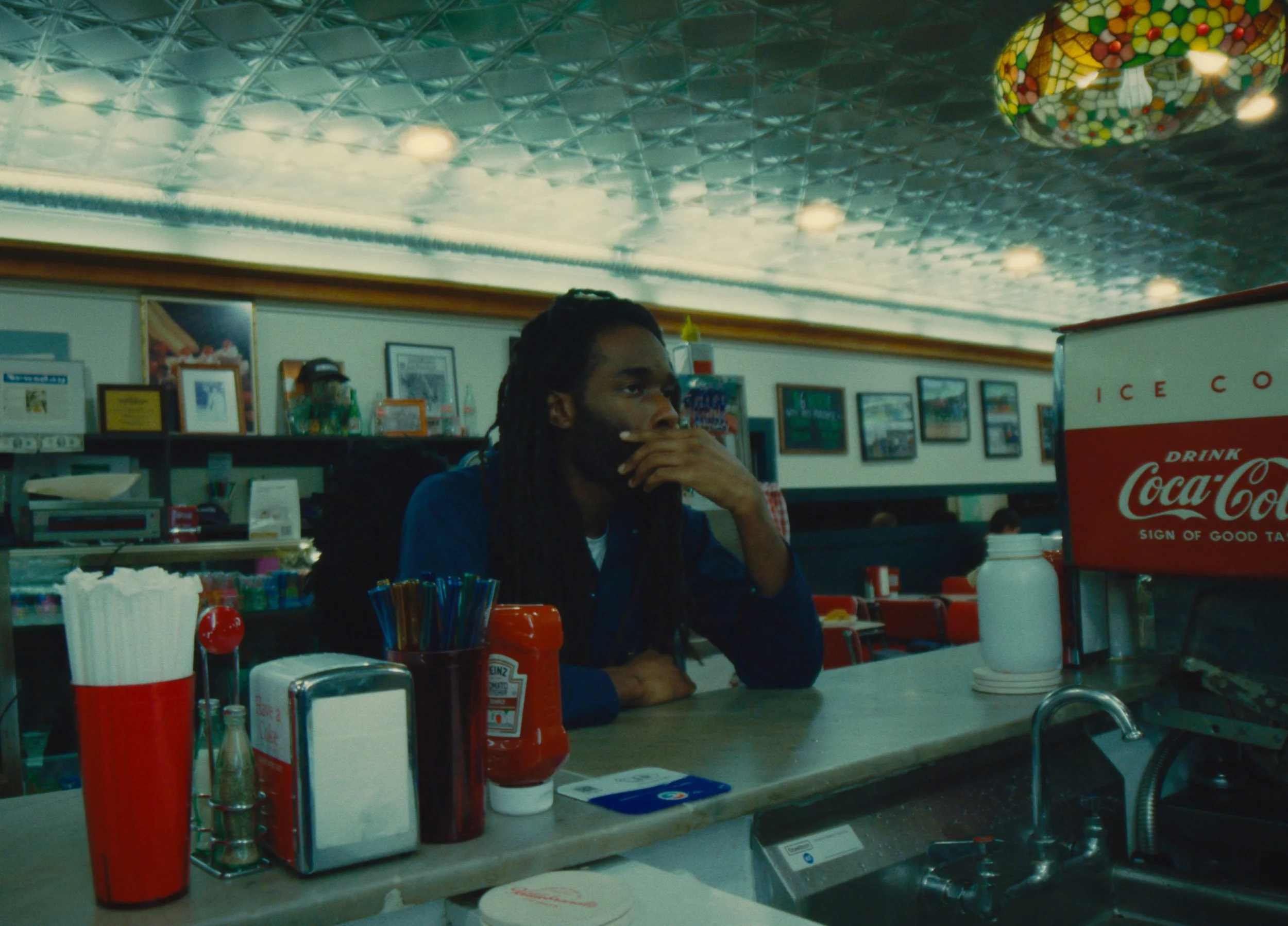 A man with long dreadlocks sitting at a counter in a diner or fast food restaurant, appearing to be waiting or deep in thought, with a Coke machine on the right and various condiments on the counter.
