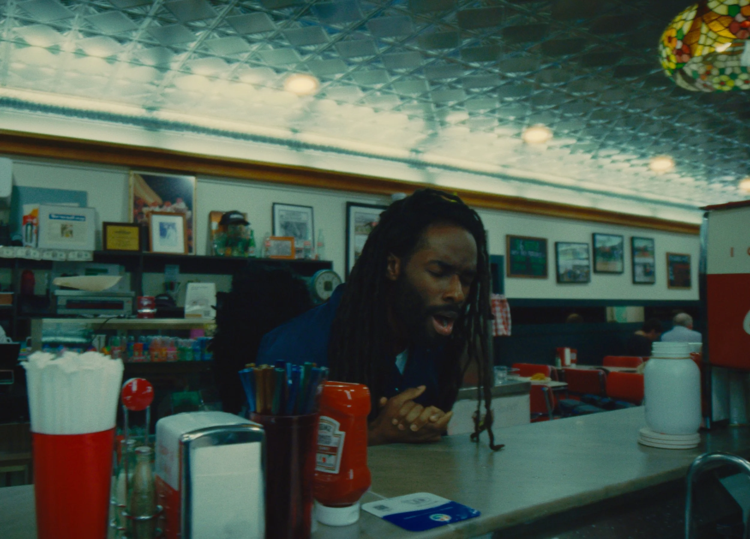 A man with long dreadlocks and a beard sitting at a counter in a diner, appearing to be singing or speaking passionately, with condiments and utensils on the counter, and framed pictures on the wall in the background.