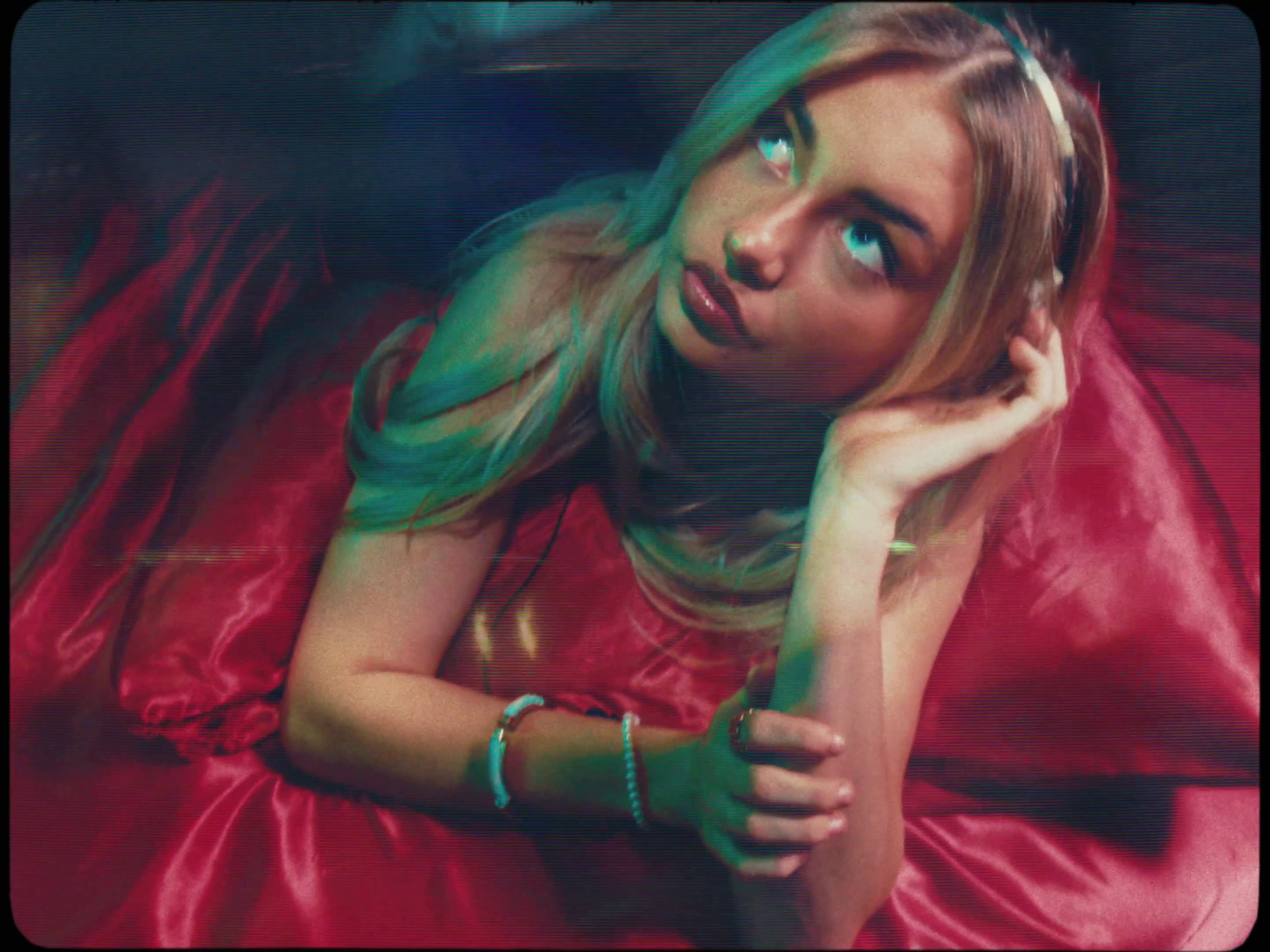 A young woman with blonde hair, wearing headphones and a red satin dress, is laying on a red satin surface, looking upward with a relaxed and contemplative expression.