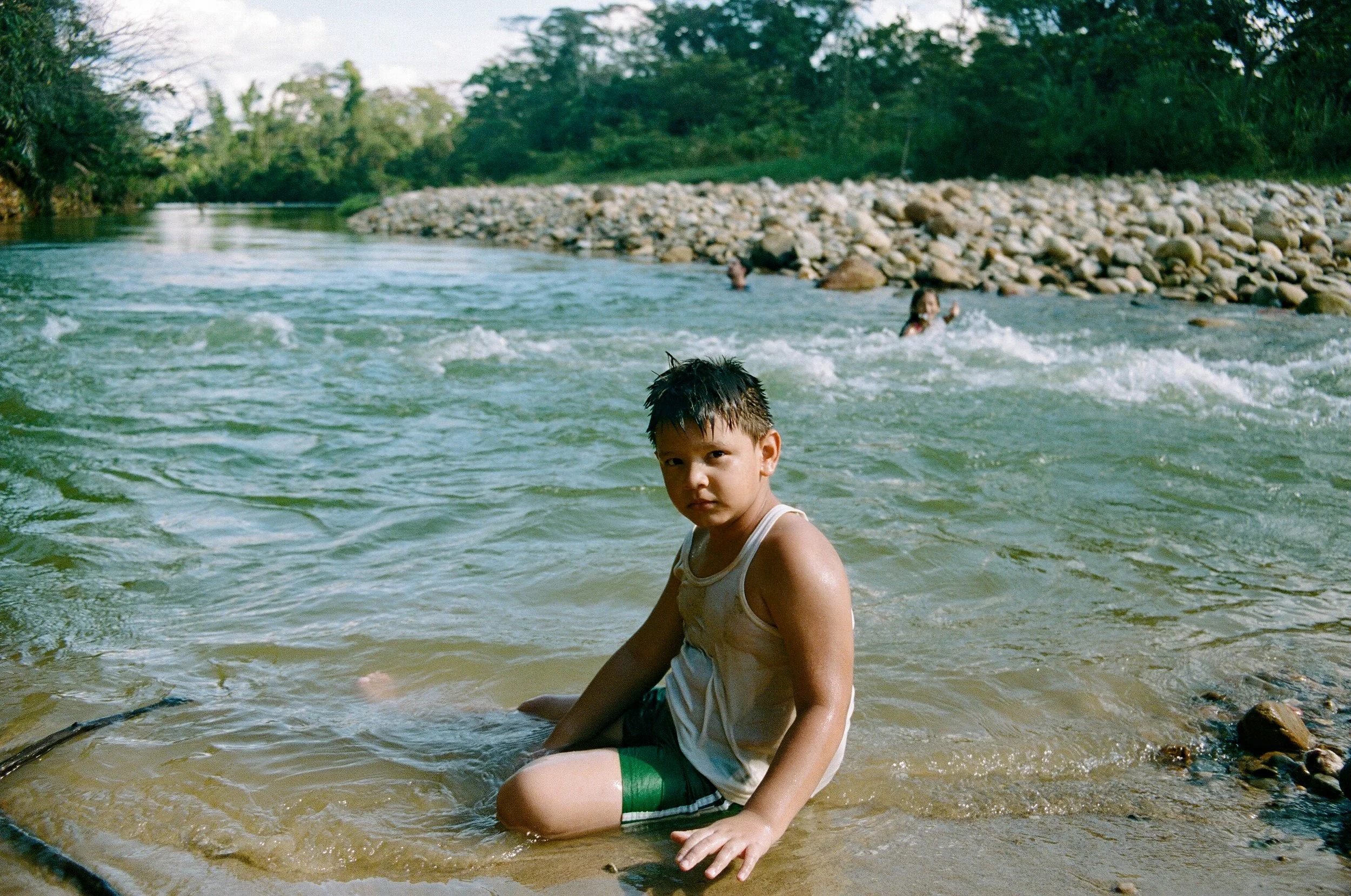 A young boy sitting in a river, wearing a sleeveless shirt and shorts, with other children playing in the water in the background, surrounded by a natural outdoor setting with trees and rocks.