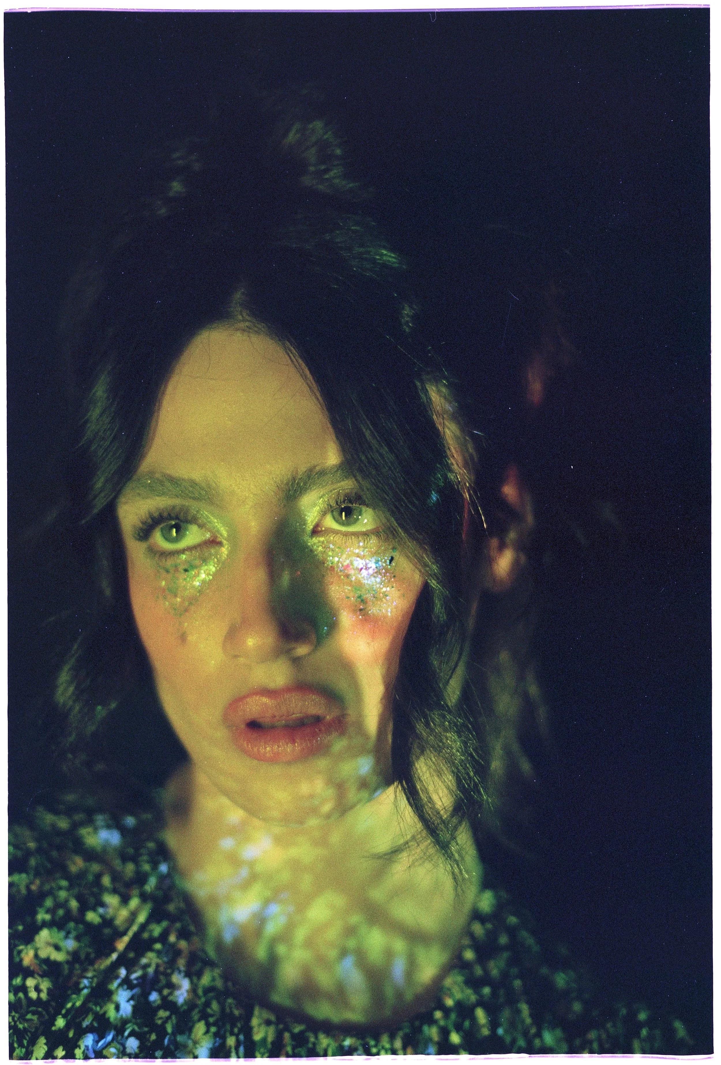 A woman with dark hair, green eyes, and glitter on her cheeks, illuminated by colorful light patterns.