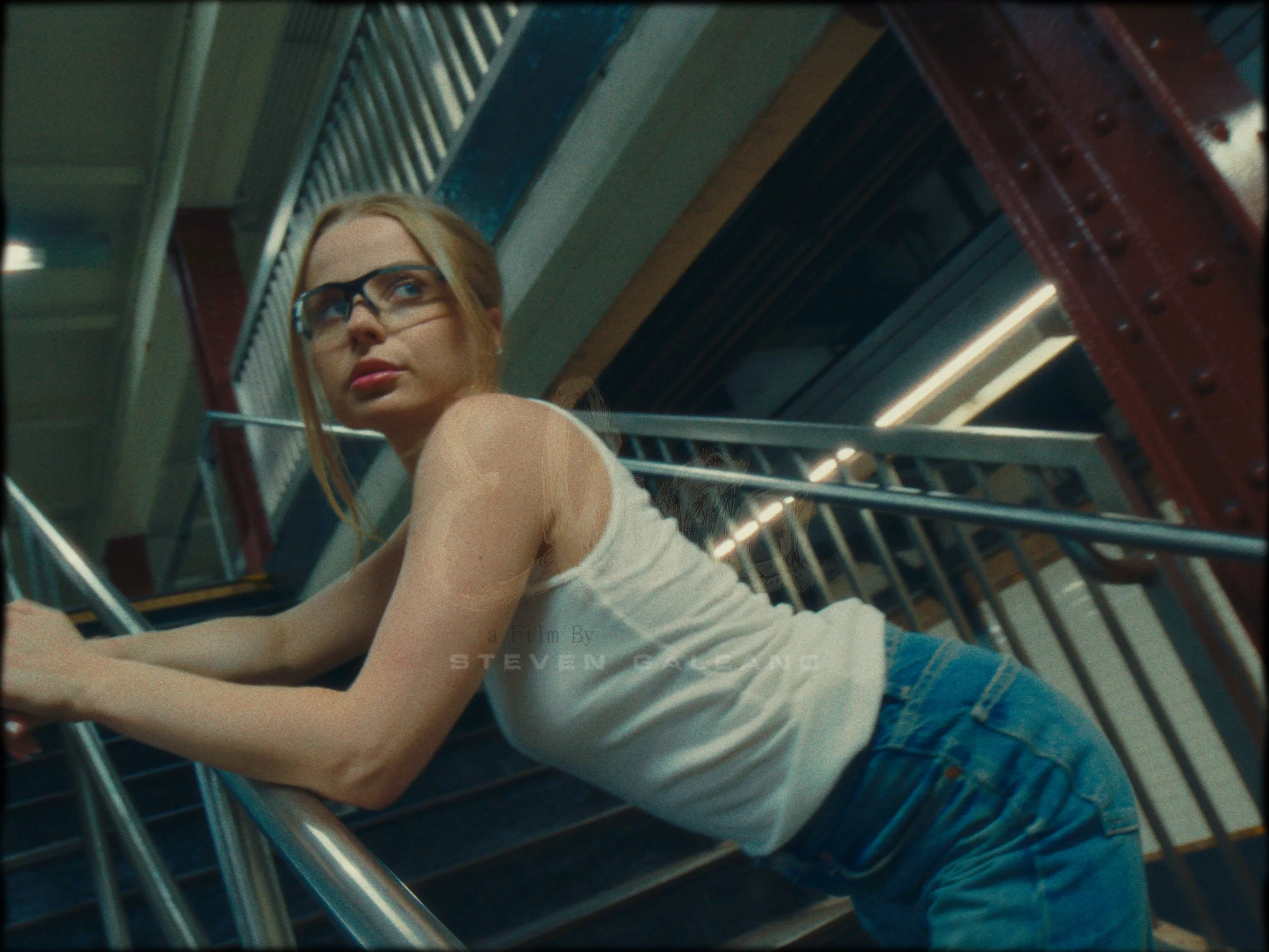 Young woman with blonde hair wearing glasses, a white sleeveless shirt, and blue jeans, leaning against metal handrails on a staircase in an industrial or warehouse-like setting.