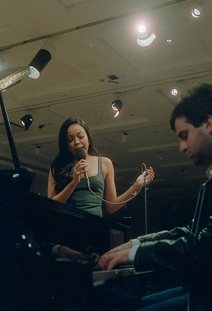 A woman singing into a microphone while a man plays the piano in a dimly lit room.