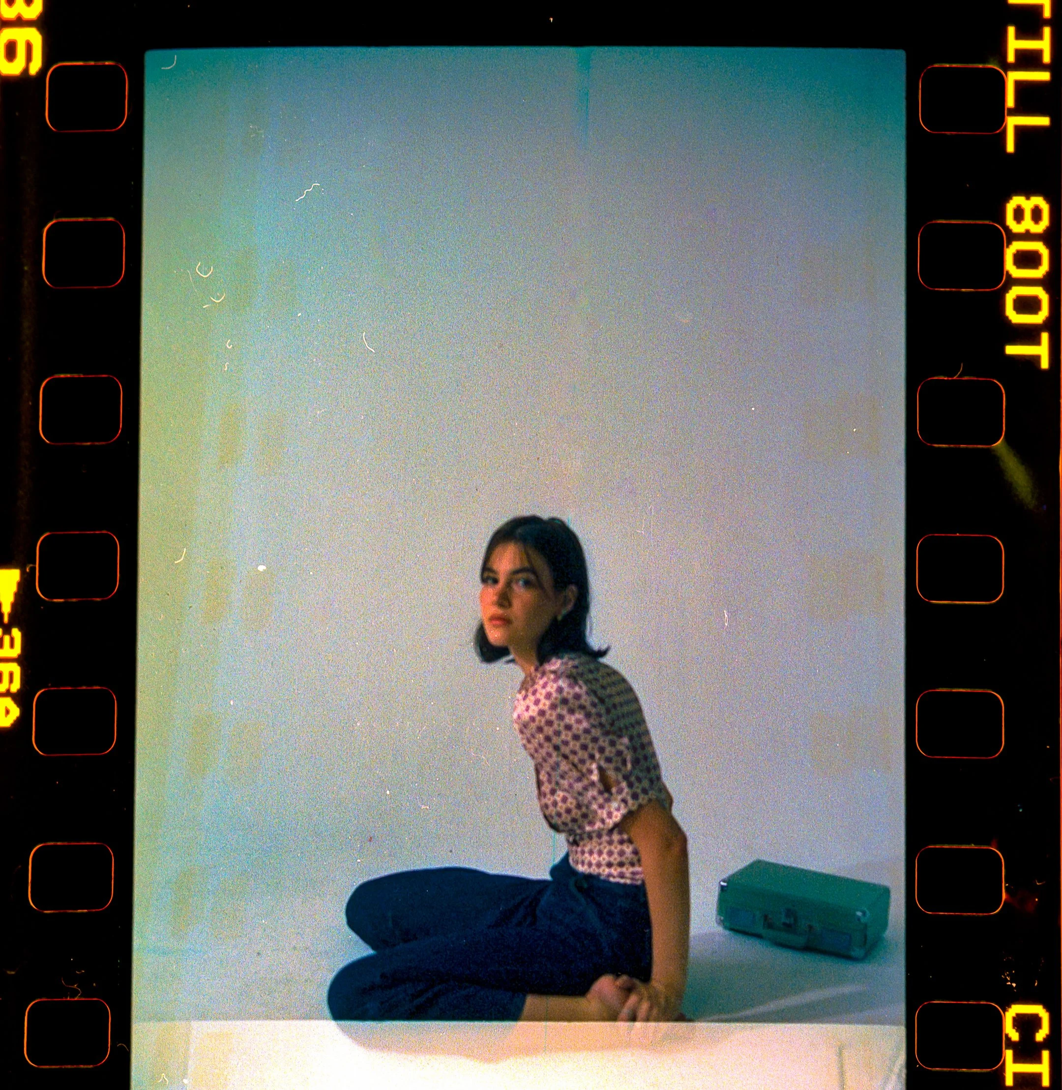 Young woman with dark hair, sitting cross-legged on the floor in front of a gradient background, wearing a patterned shirt and dark pants. There is a small teal-colored box next to her.