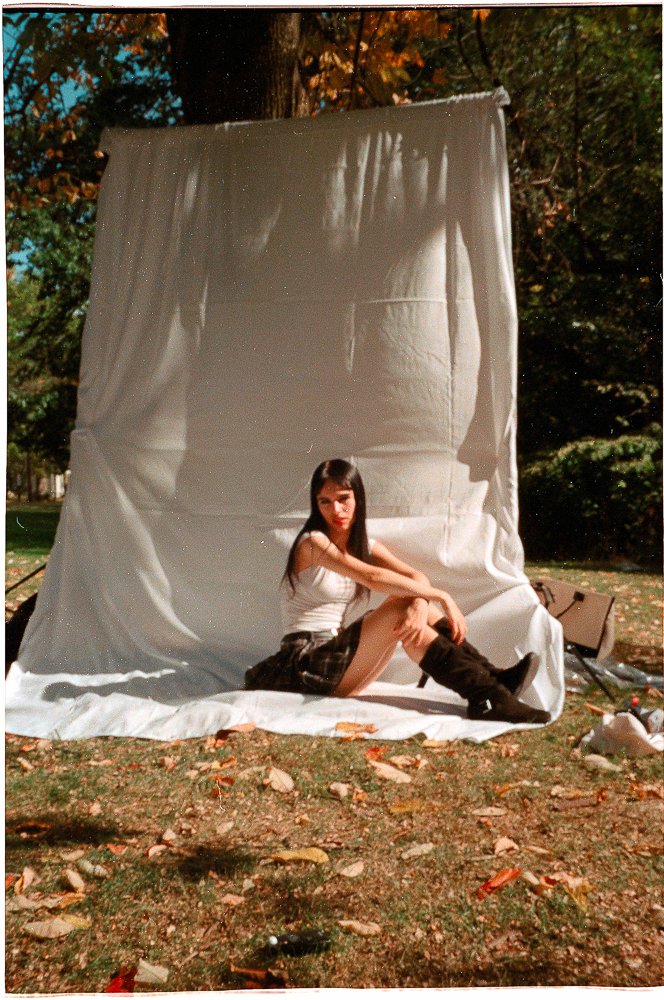 A young woman sitting on the ground in front of a white fabric backdrop in an outdoor park setting.