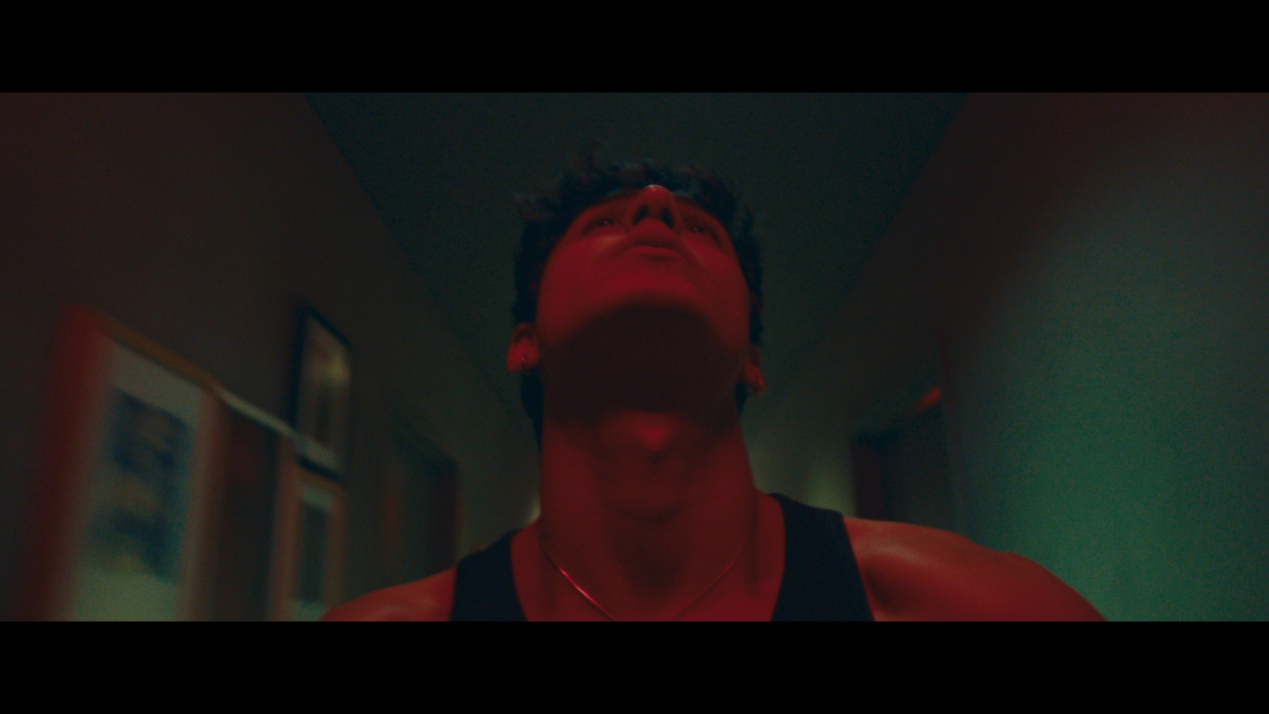 A person with short dark hair, wearing a black tank top and a silver necklace, looking upward with a red light cast on their face, in a dimly lit room.