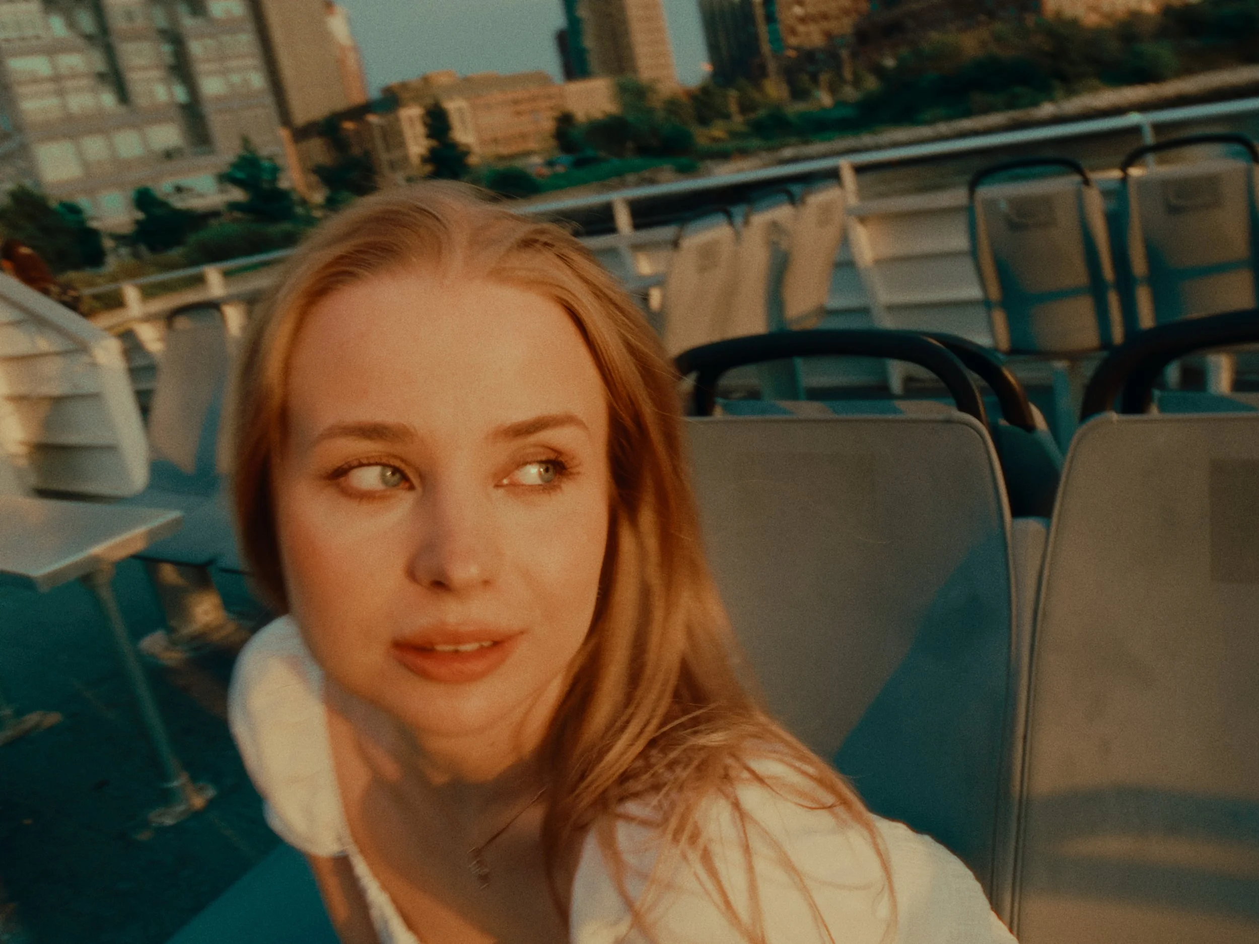 A young woman with red hair and blue eyes, looking to her right, on an outdoor ferry with empty seats and city buildings in the background during sunset.