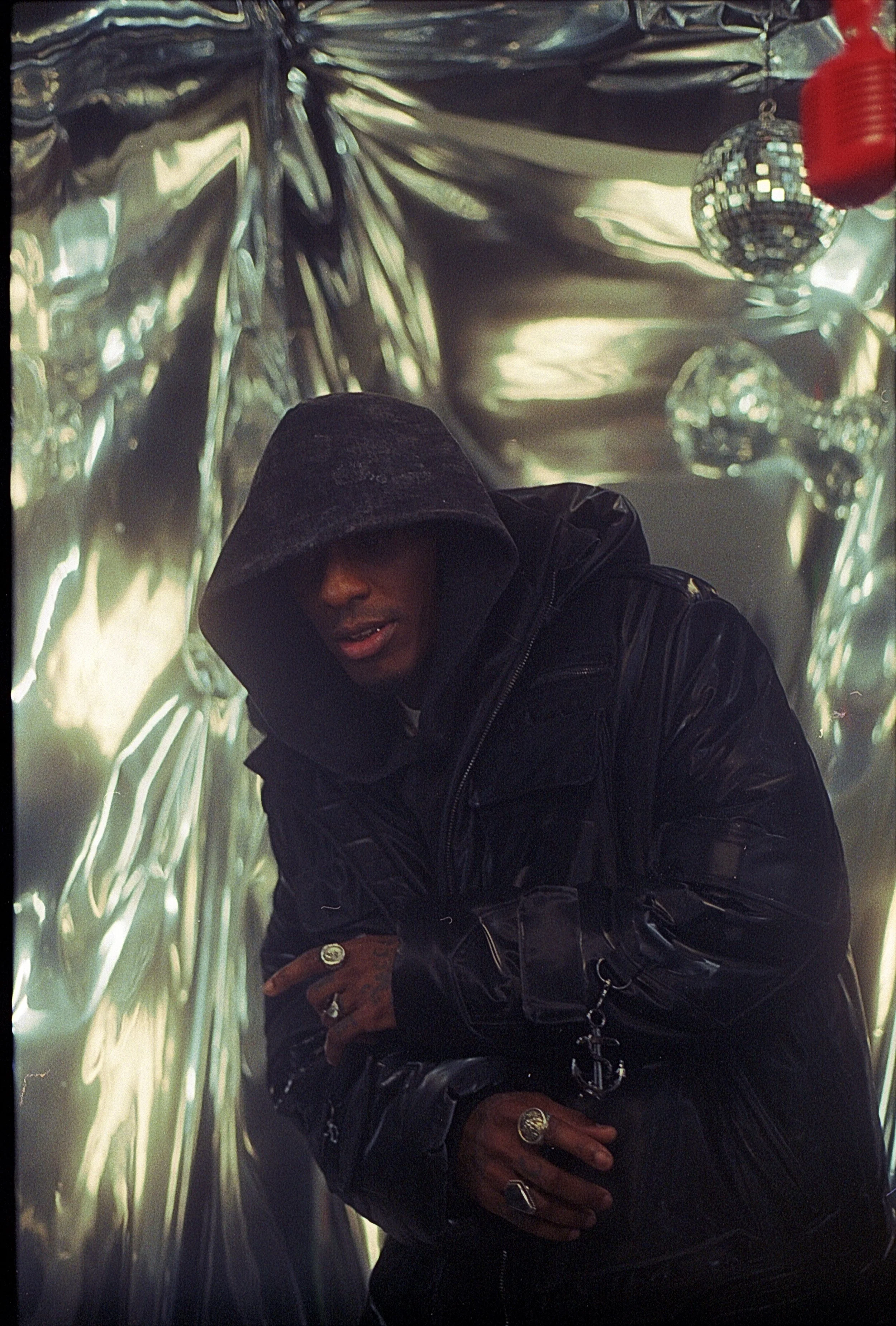 A person wearing a black hooded jacket with the hood over their head, standing in front of a reflective metallic background with disco balls and a red object. The person's face is partially visible, with their mouth slightly open and hands adorned with rings.