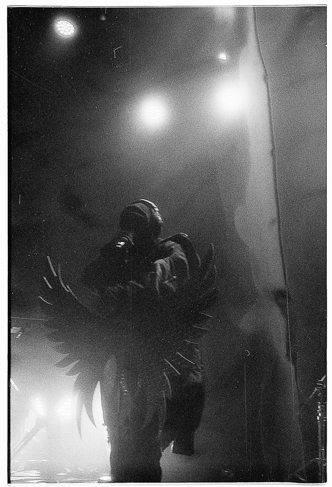 A person wearing dark armor with wings, standing in a dimly lit environment with bright lights and reflecting surfaces.